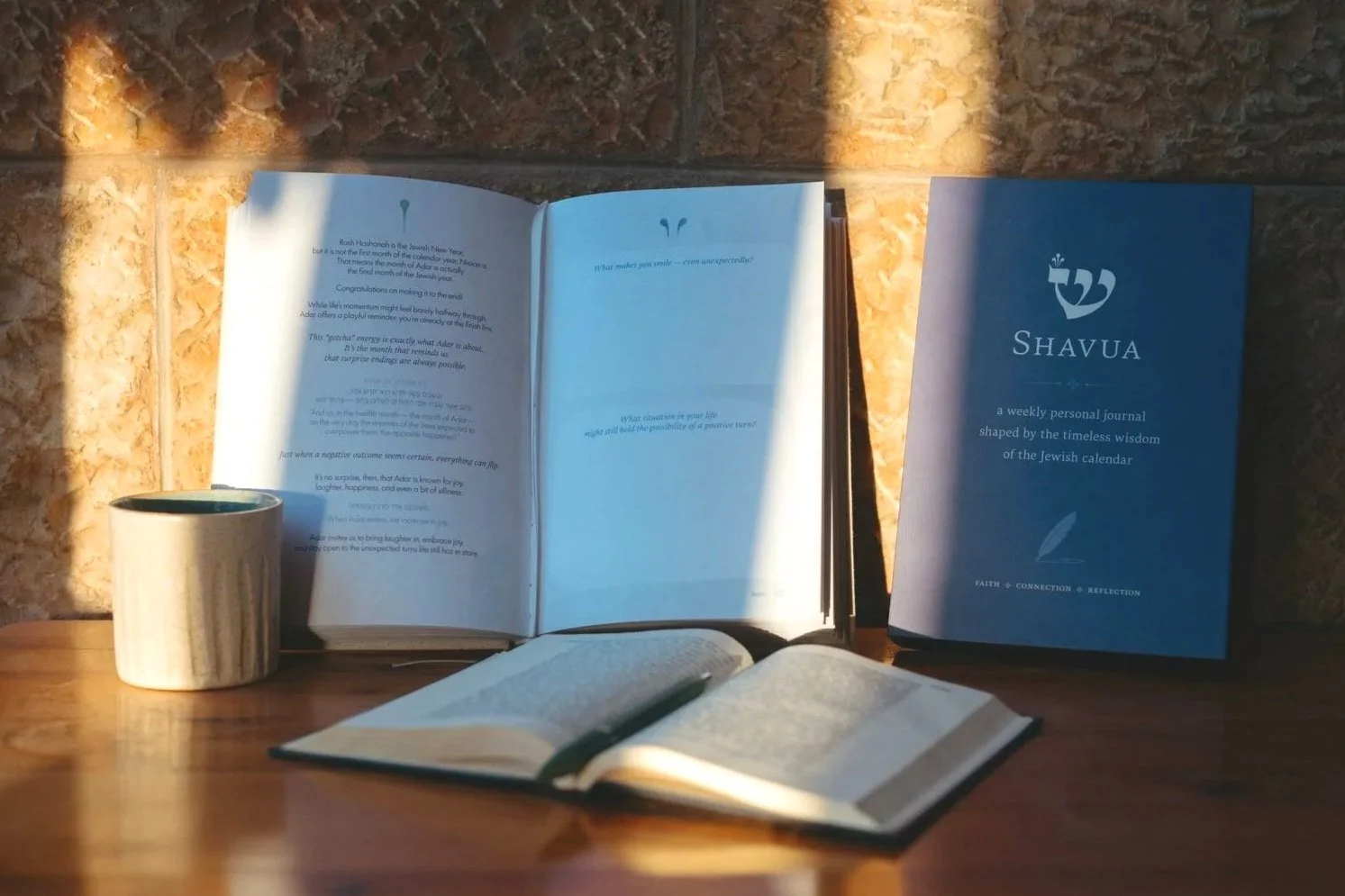 Open books, a cup of coffee, and a closed book titled 'Shavua' on a wooden table with a brick wall in the background, illuminated by warm sunlight.