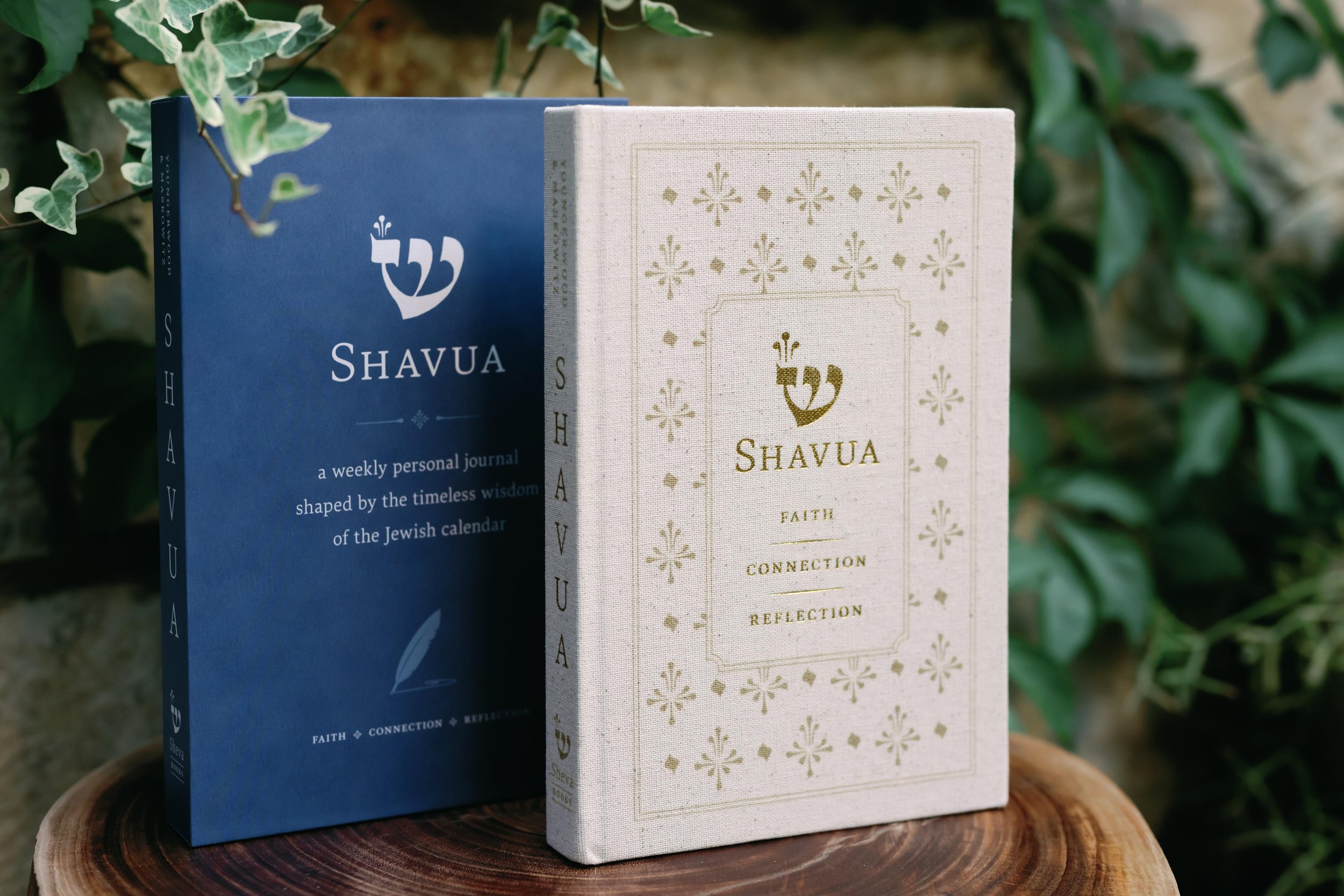 Two books titled 'Shavua', one with a blue cover and one with a beige cover, standing upright on a wooden surface, with green leafy plants in the background.