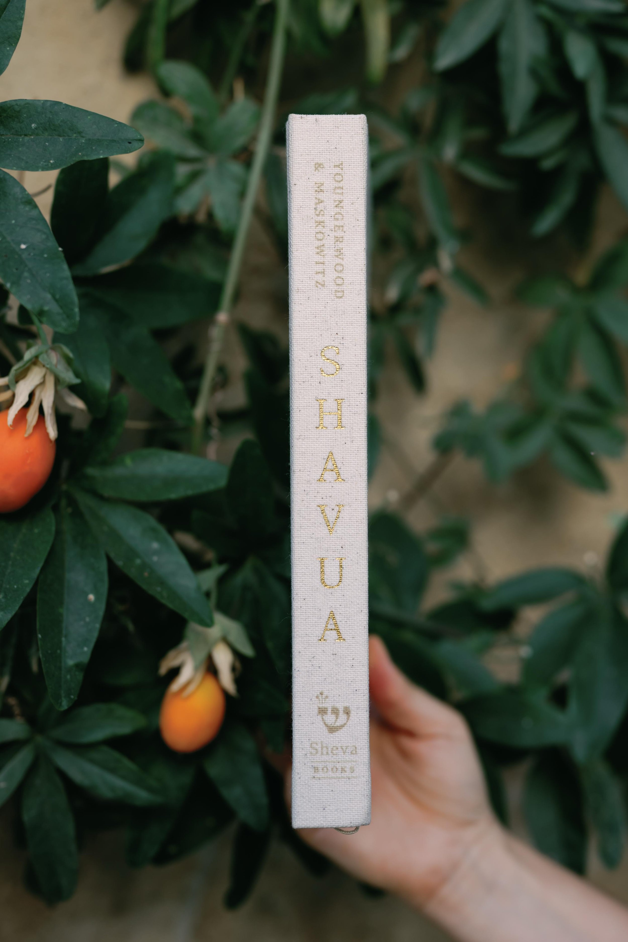 A hand holding a white fabric book with gold lettering that reads "SHAVA" among green shrubbery with orange fruit visible.