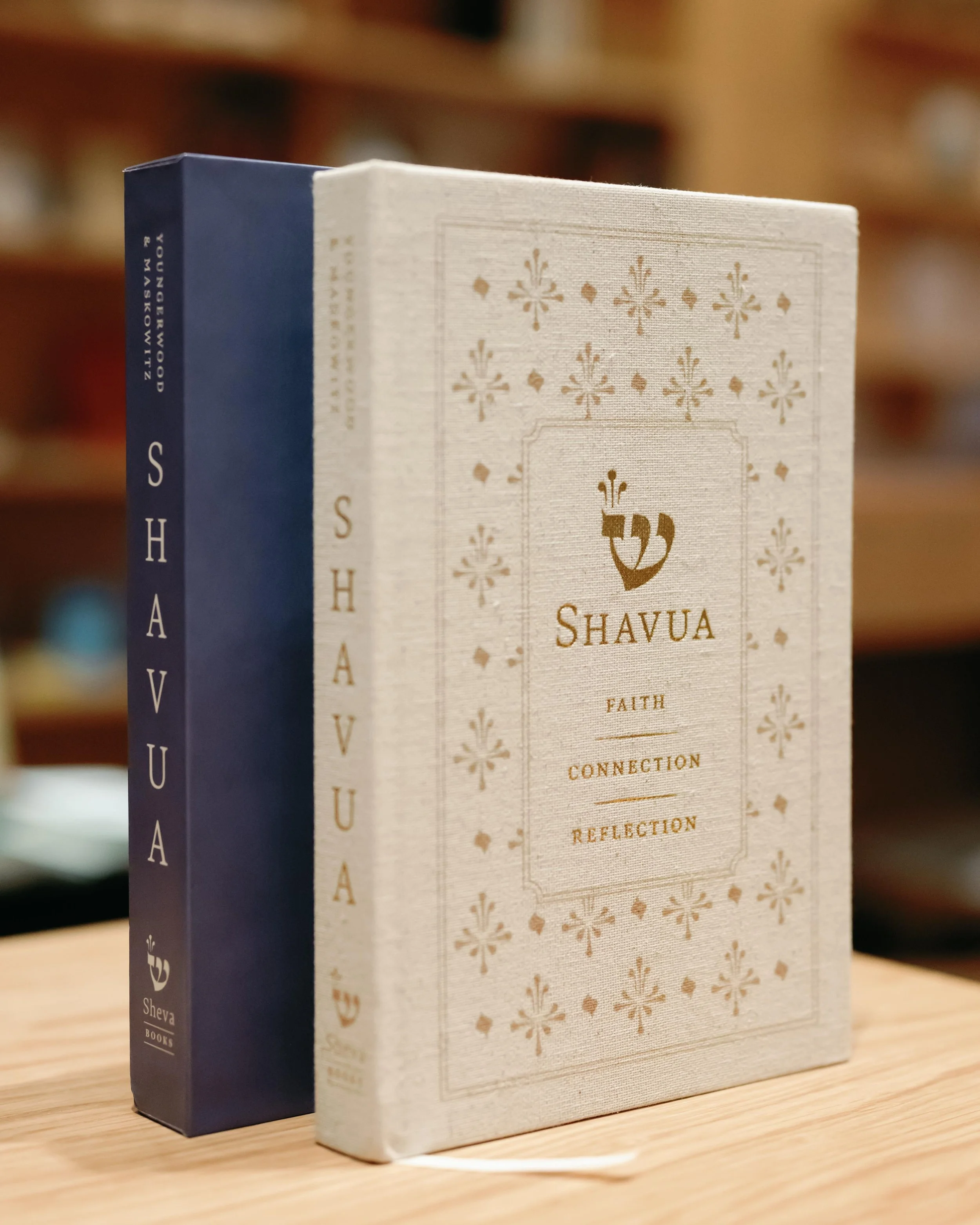 A beige hardcover book titled "Shavuot" with topics such as faith, connection, and reflection, standing upright next to a dark blue hardcover book on a light wooden surface.
