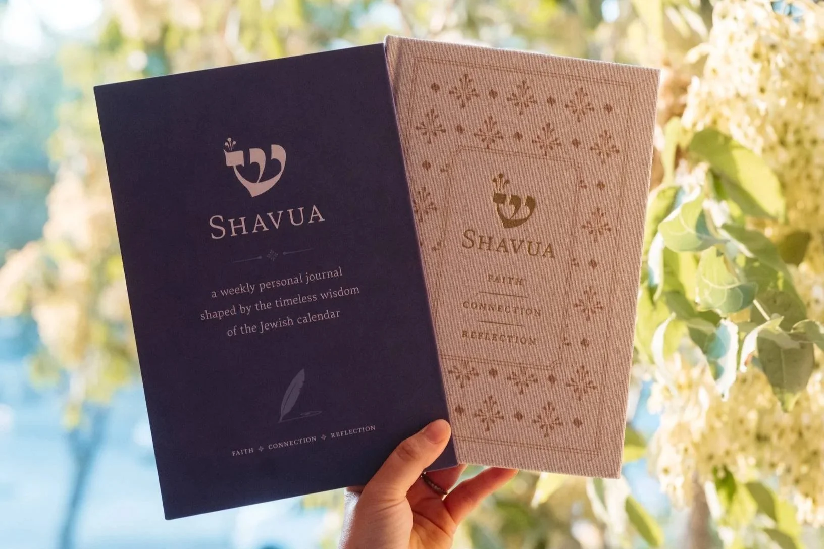 Two holding up books titled Shavua, one in blue and one in light pink, with text describing a weekly personal journal based on the Jewish calendar, against a background of green foliage and sunlight.