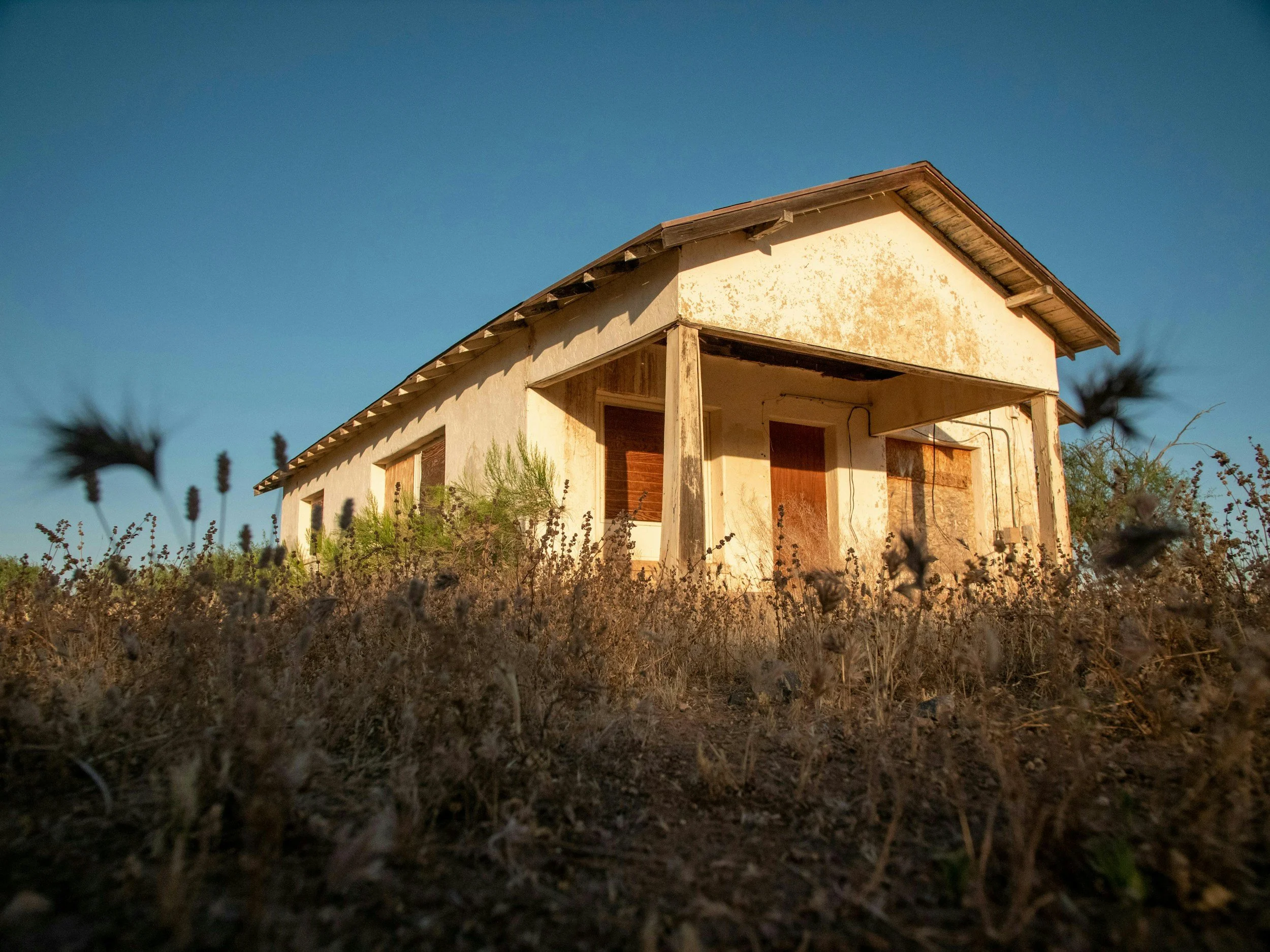 An exterior view of an unfinished house with exposed wood beams, boarded-up windows, and peeling paint, set against a clear blue sky. Tall grass and weeds grow in the foreground.