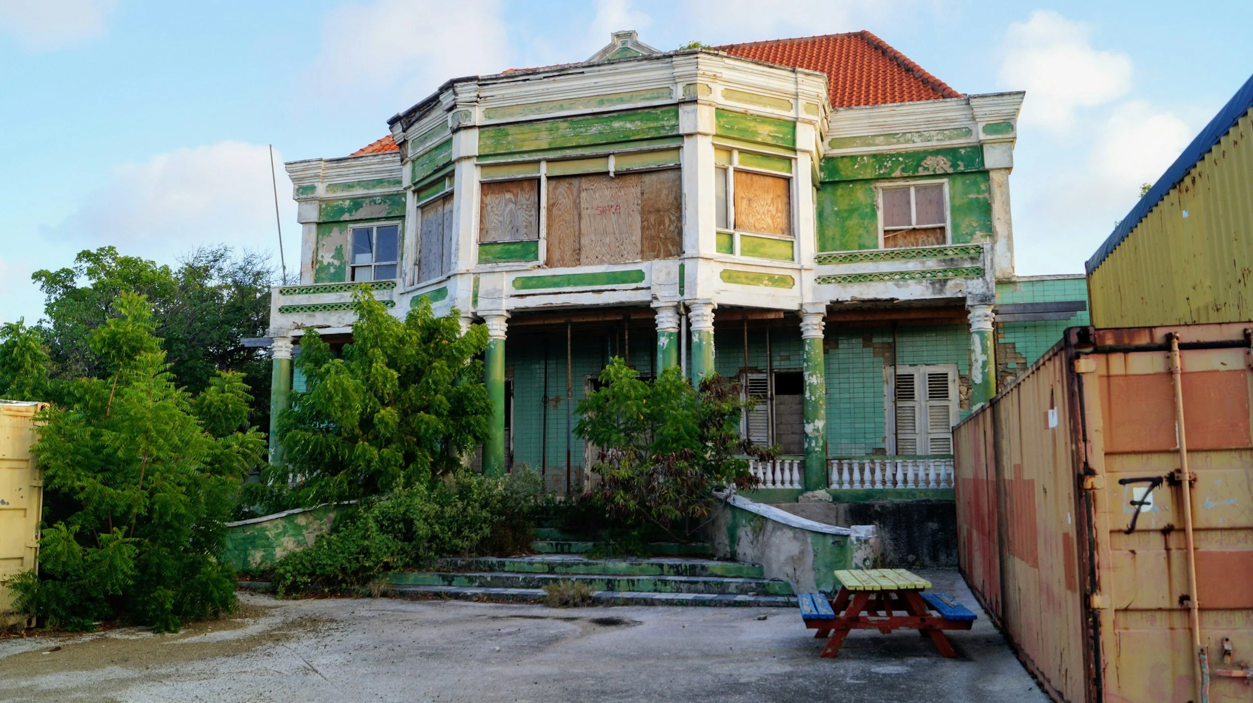 Abandoned green and white house with boarded-up windows, surrounded by trees and shipping containers.