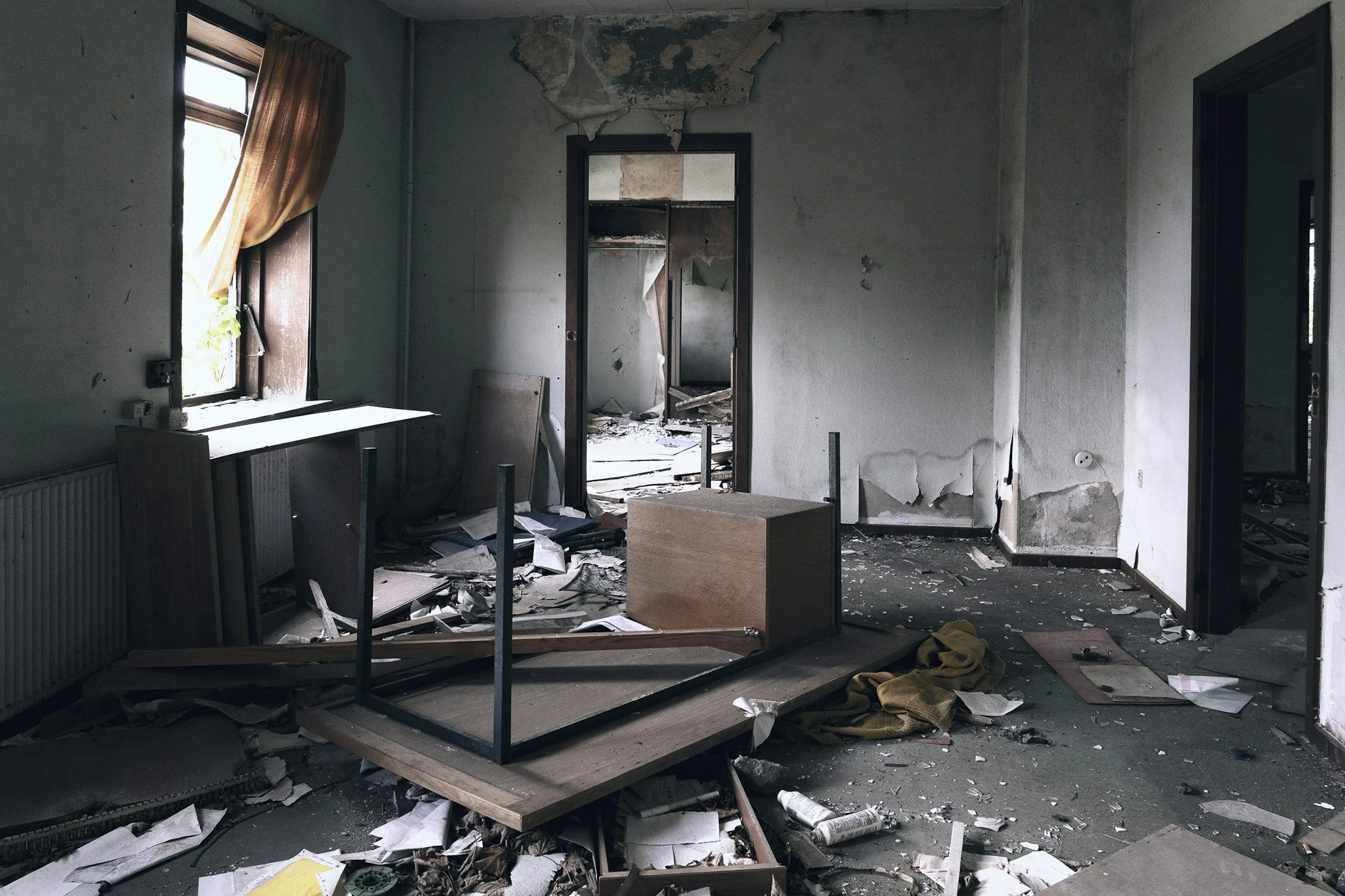 A room in disrepair with peeling paint and damaged walls. The floor is cluttered with debris, papers, and broken furniture. A window has a torn curtain, and a mirror on the wall reflects the chaotic scene.