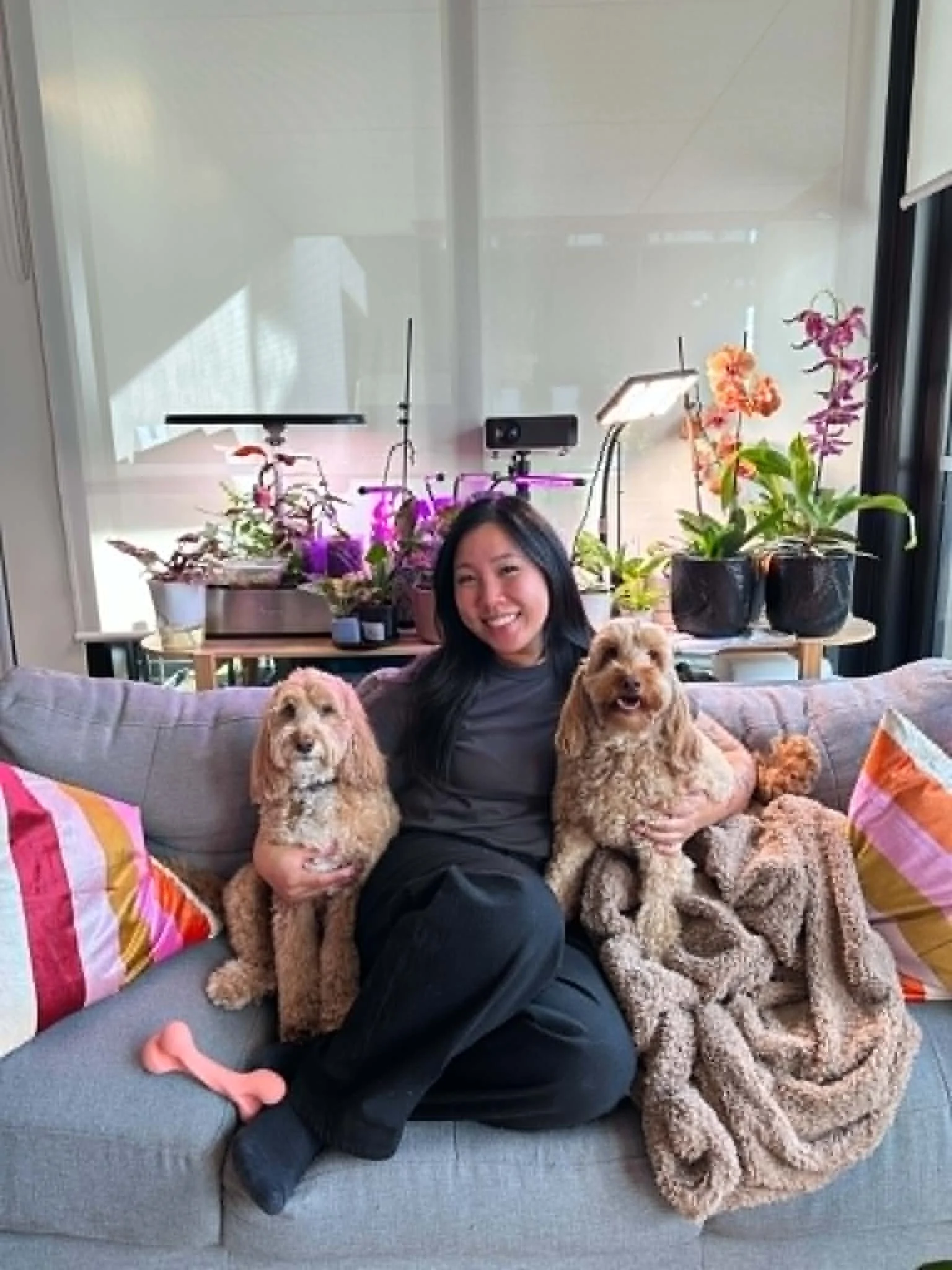 Pet sitter sitting on couch with the dogs she pet sits for with a shelf of potted plants behind her at her home