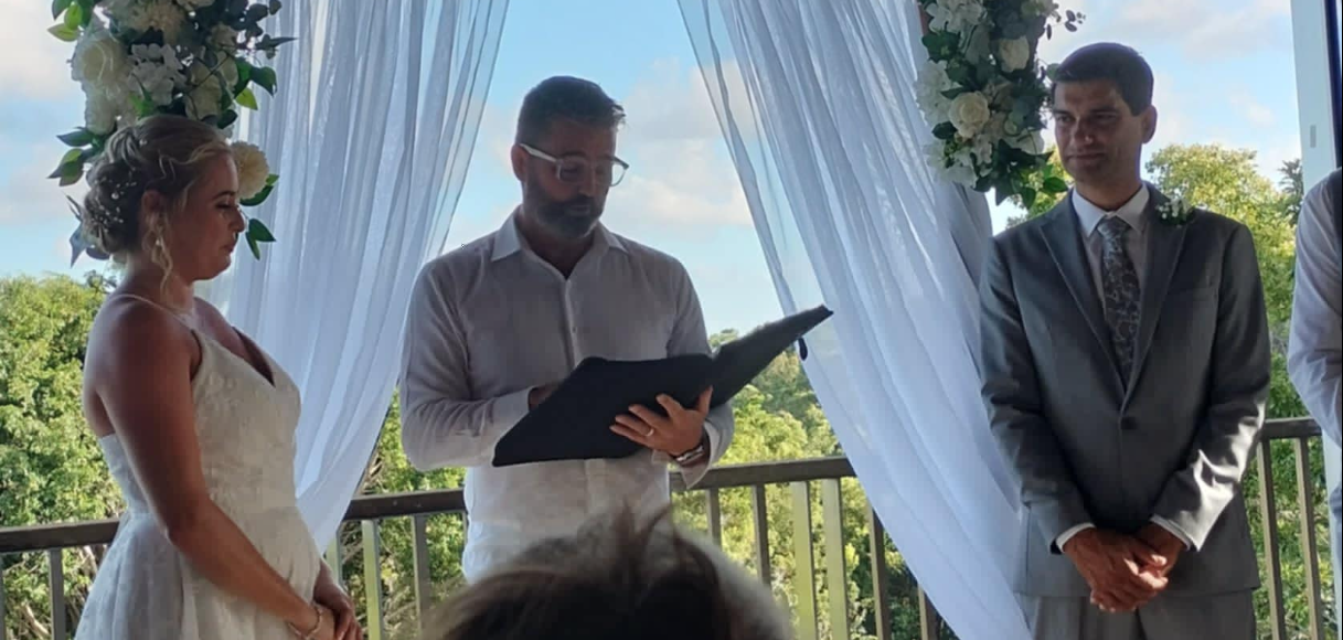 A bride and groom stand under a decorated arch, during a wedding ceremony outdoors, with an officiant reading from a book. The bride is in a white lace dress, and the groom is in a gray suit, on a balcony with green trees in the background.