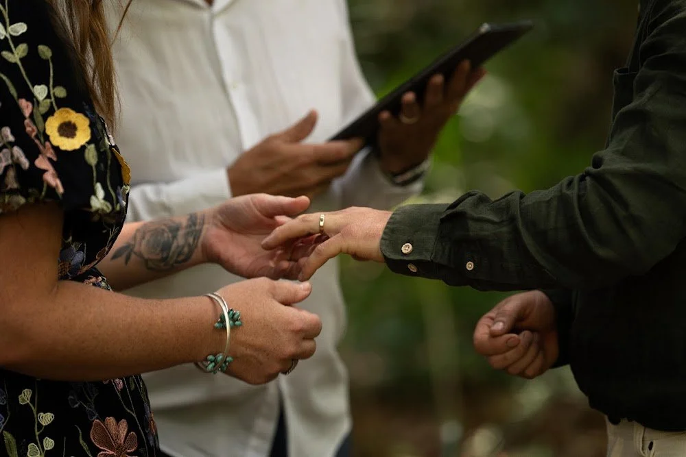 A couple holding hands in a forest while a person in the background reads from a tablet.