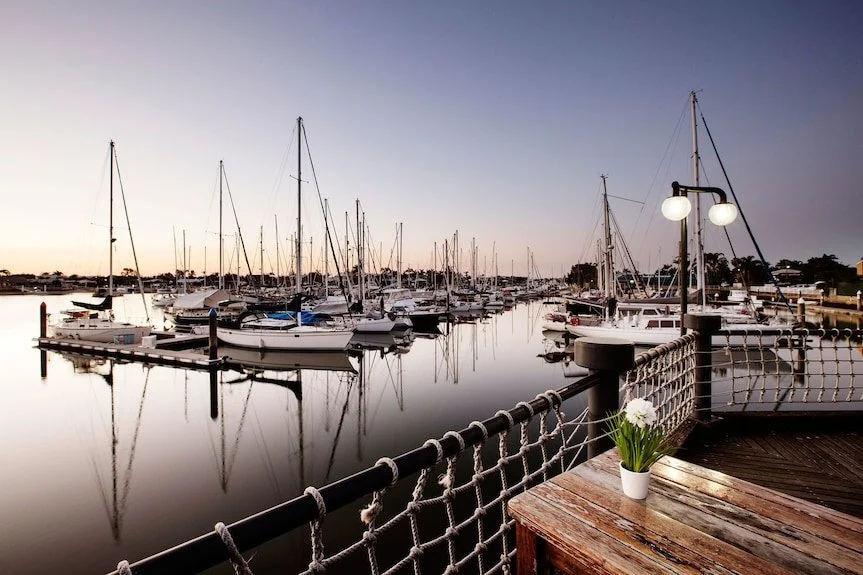 Yacht harbor with boats moored in calm water at dusk, wooden deck with potted plant, and outdoor lighting.