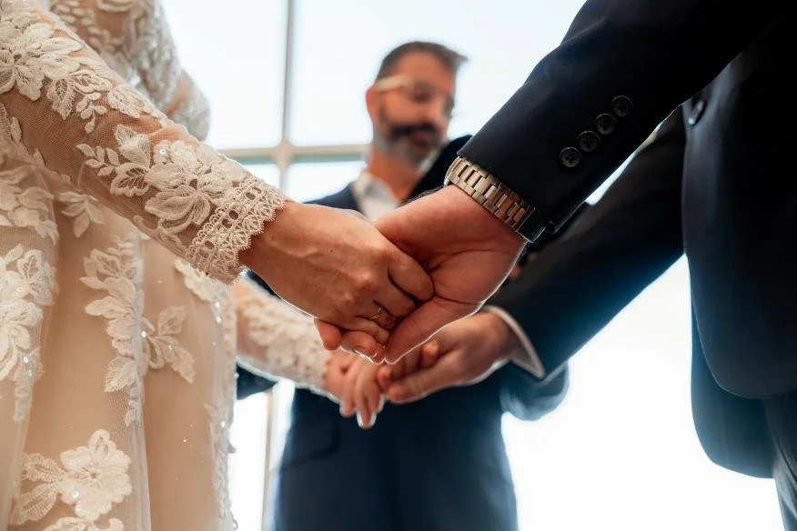 Close-up of a bride and groom holding hands during a wedding ceremony, with an officiant in the background.