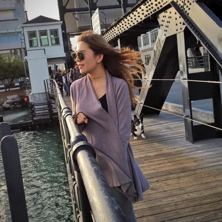 A woman with long hair wearing sunglasses, a purple coat, and black top, standing on a wooden pier with a black and white dog behind her, near a waterway and a bridge.
