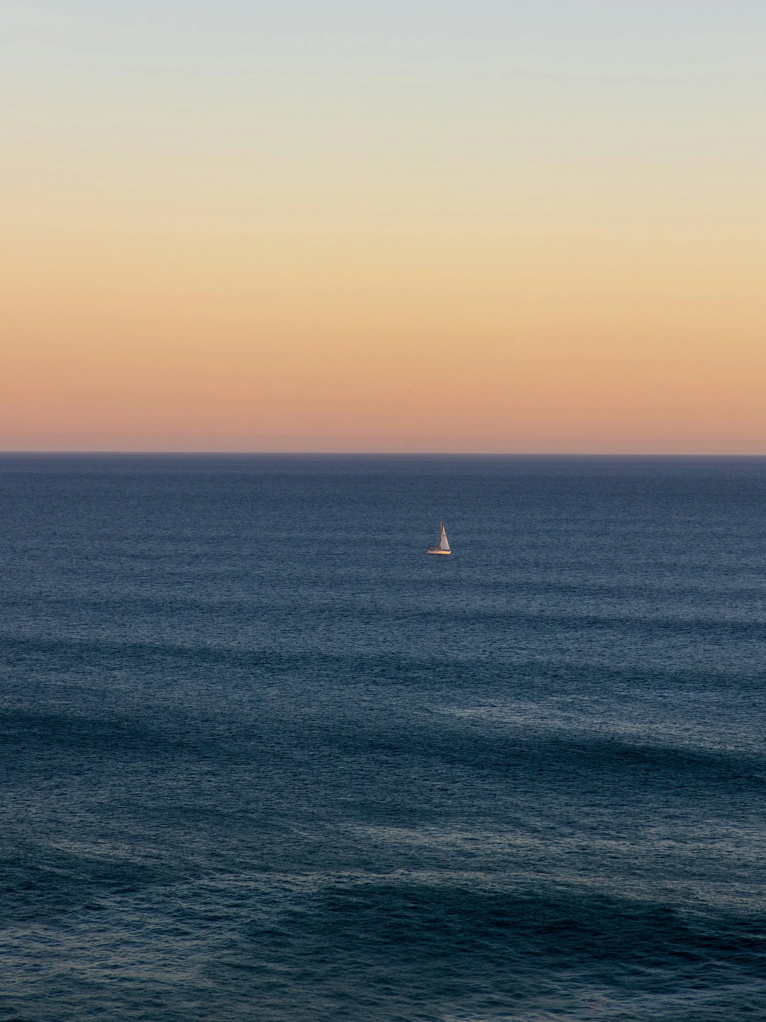 A single sailboat on calm ocean waters during sunset with a pastel gradient sky.
