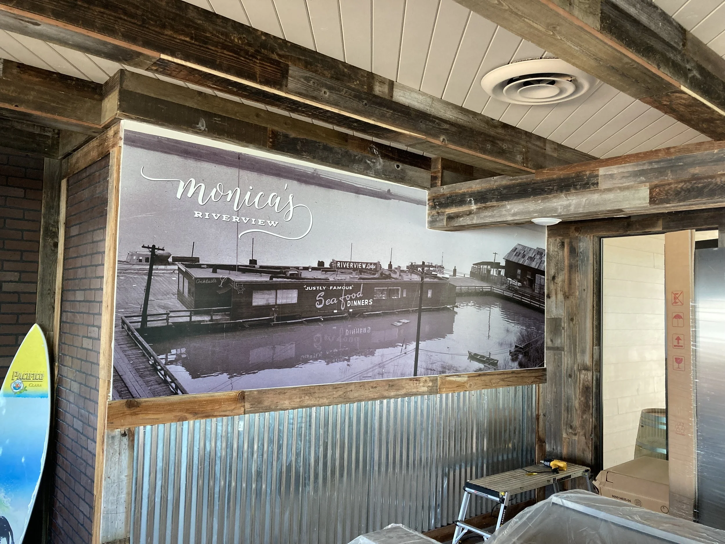 Interior of a building with wooden beams and a corrugated metal wall, featuring a large black and white mural of a waterfront scene with buildings and a sign that reads 'Monica's Riverview' and 'Seafood Dinners'.