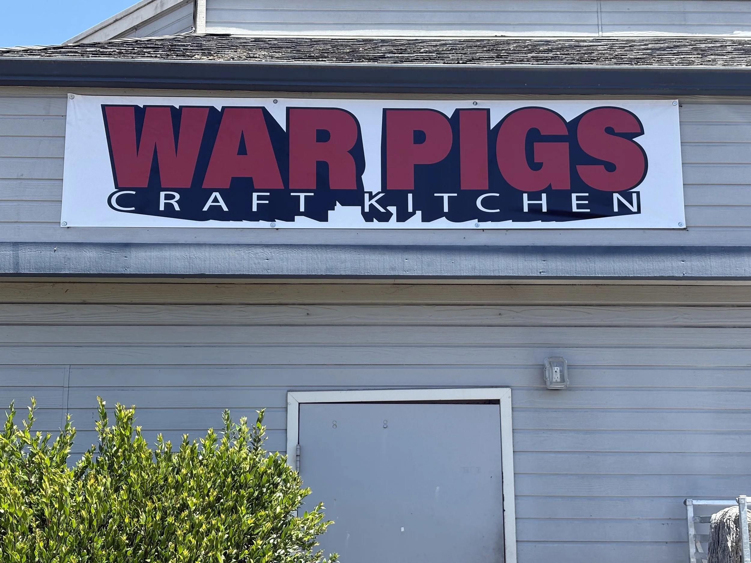 Sign on a building that reads "WAR PIGS CRAFT KITCHEN" with beige siding and a part of a bush in foreground.