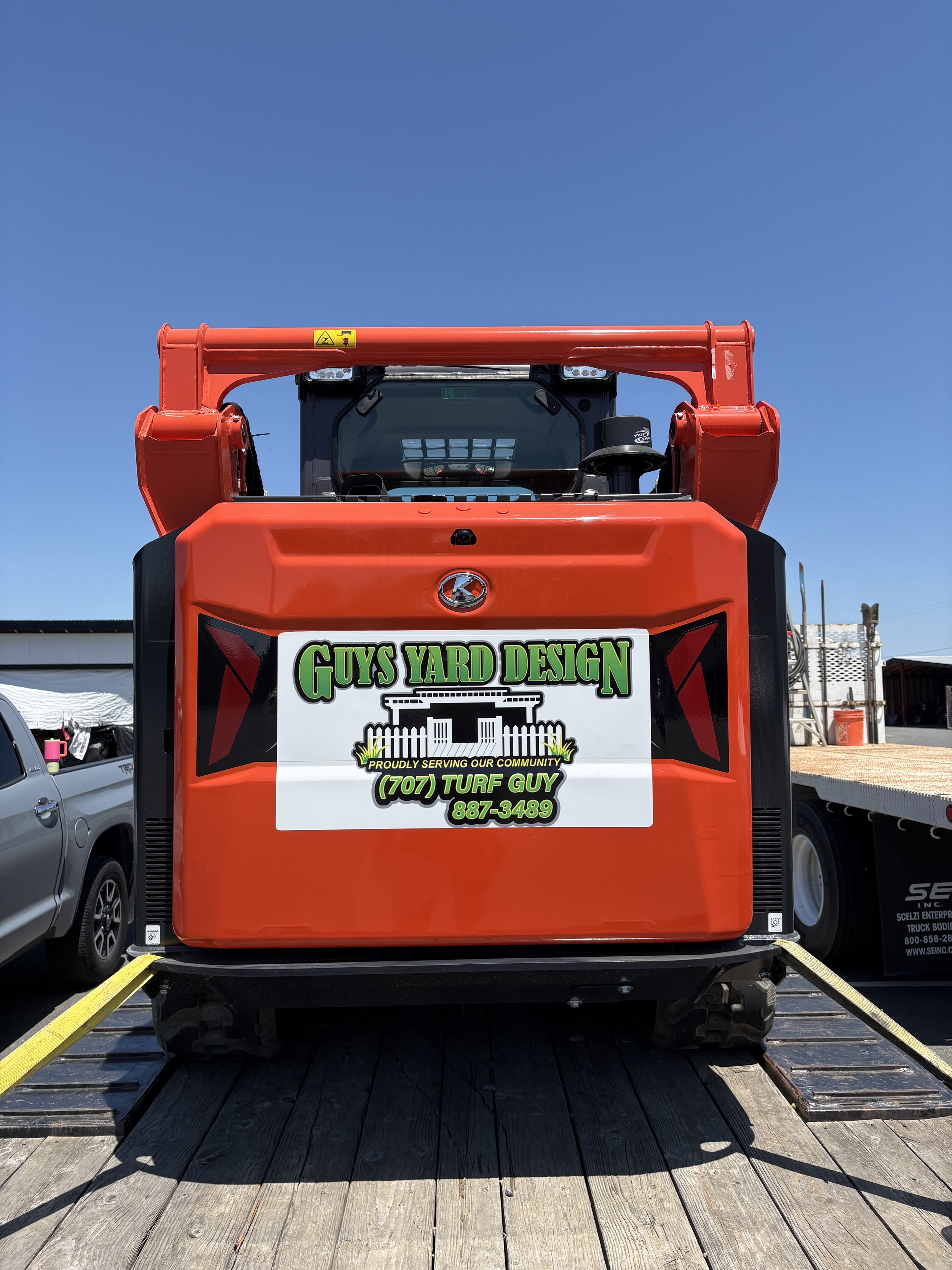 Front of a red utility truck with a sign that reads 'Guys Yard Design, Proudly Serving Our Community, (707) Turf Guy, 887-3489,' parked on a wooden dock under a clear blue sky.