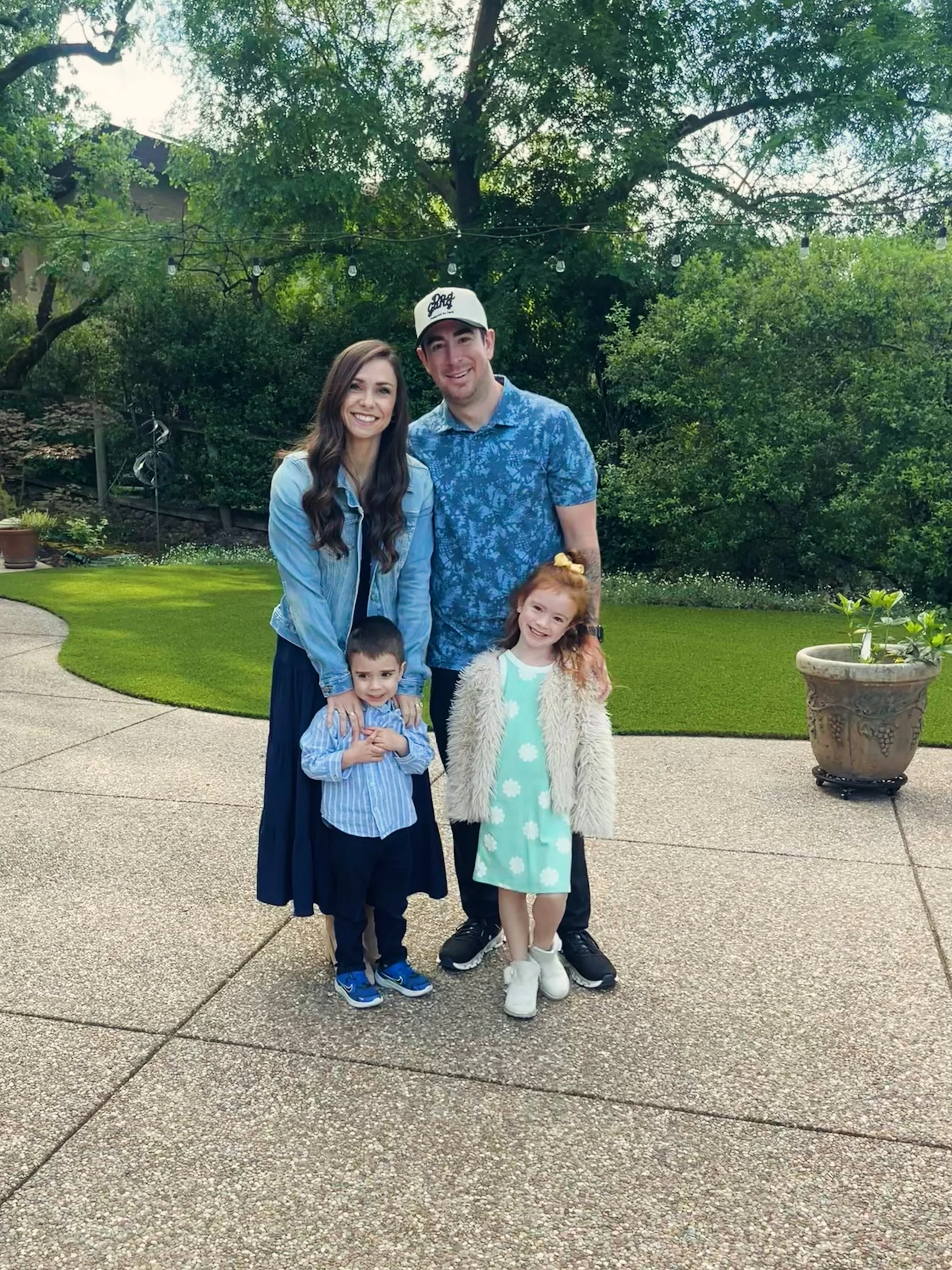 A smiling family of five, including a woman, a man, two young children, and a baby, standing outdoors on a concrete patio surrounded by trees and greenery. The woman has long wavy brown hair, and the man is wearing a white cap. The girl has red hair and is wearing a light green dress with a cream-colored faux fur coat, and the boy has short dark hair and is wearing a blue striped shirt. A large ceramic planter with a small plant is visible to the right.