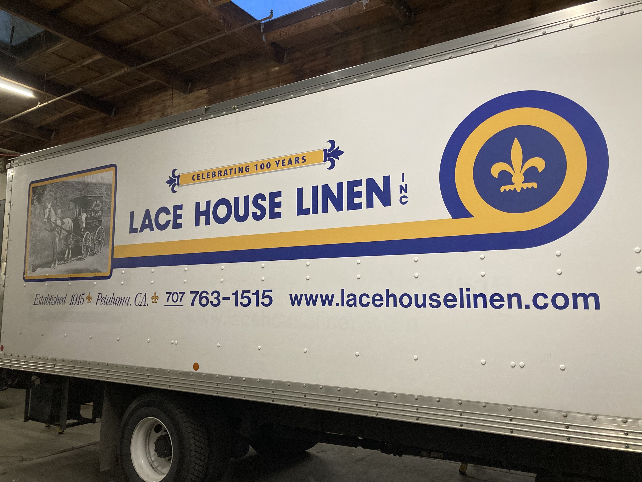 A large white truck with a banner celebrating 100 years of Lace House Linen. The banner features the company's logo with a blue and yellow design, and a historical photo of a horse-drawn carriage. The banner also displays the company's contact information and website.