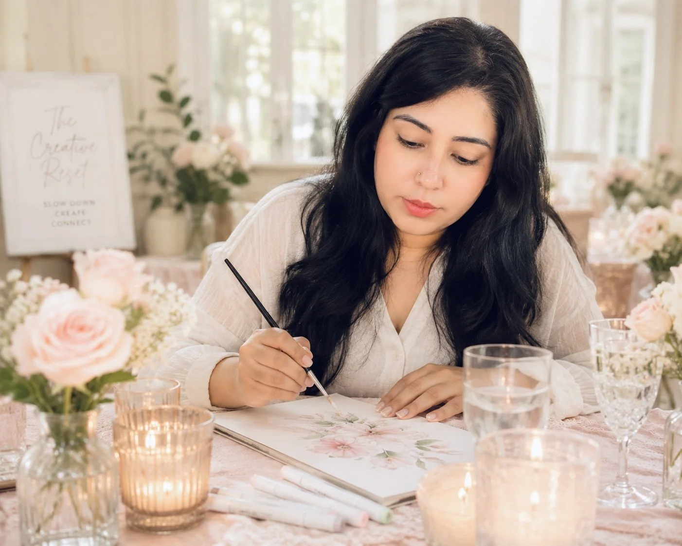 Woman with long dark hair painting a floral watercolor artwork at a craft event, surrounded by candles and pink flowers, in a bright room with large windows.