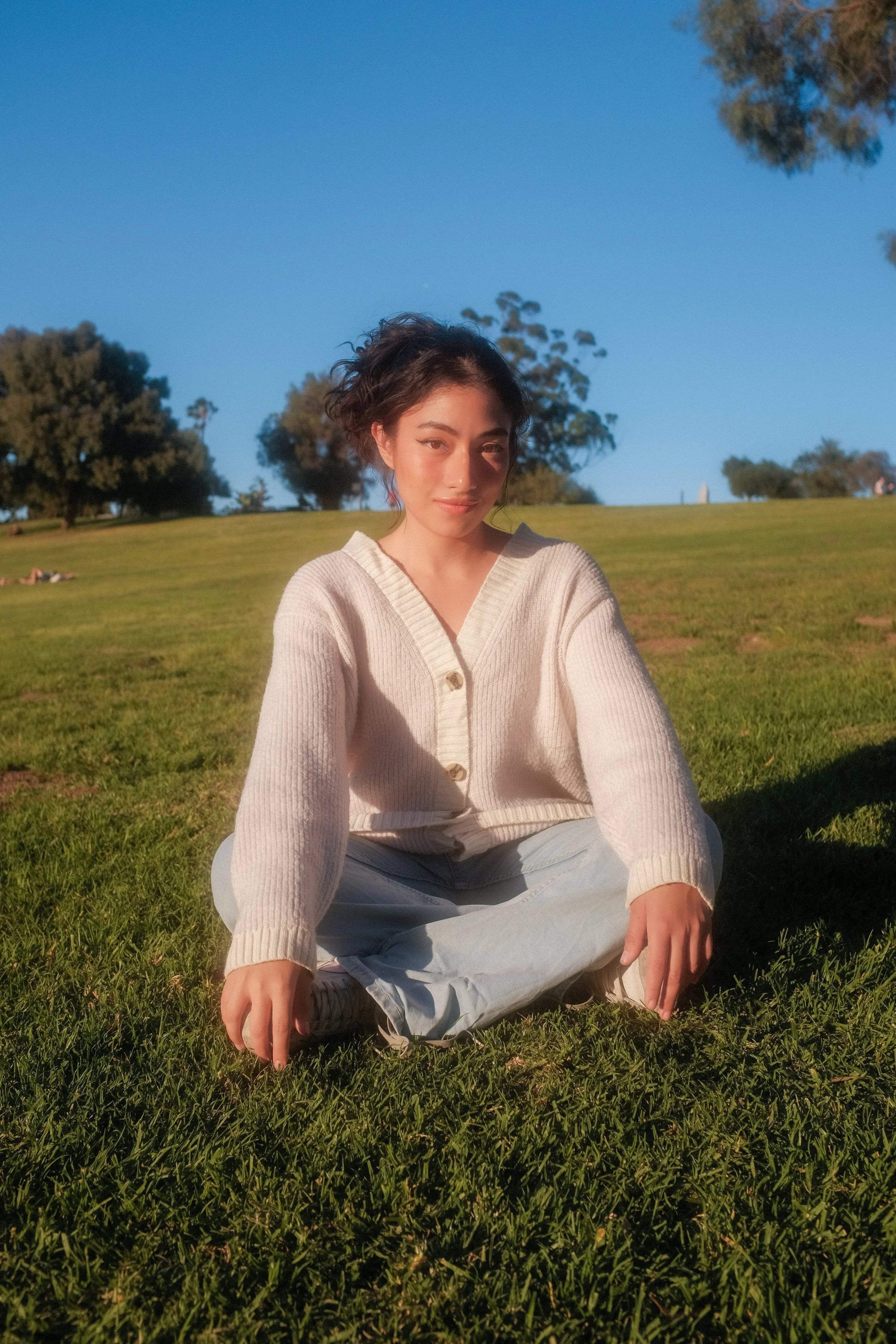 A young woman with dark hair sitting cross-legged on green grass in a park during daylight, with trees and a clear blue sky in the background.