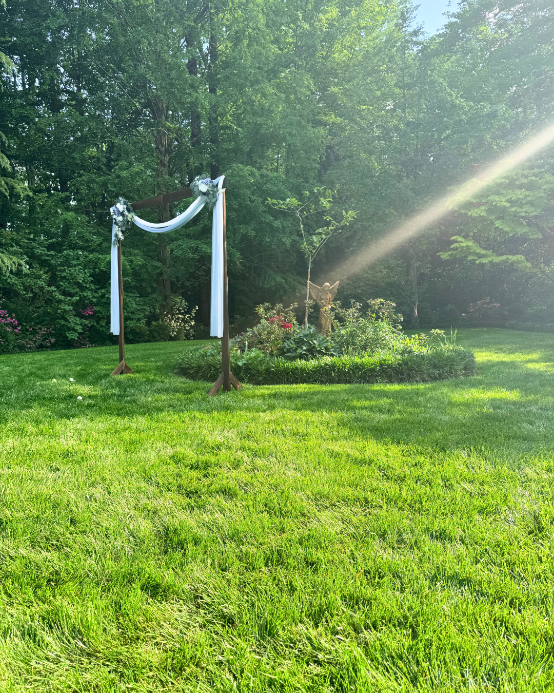 A wooden arch decorated with white fabric and flowers stands on a lush green lawn in a garden, with a statue of a dog and a small tree in the background, illuminated by sunlight.