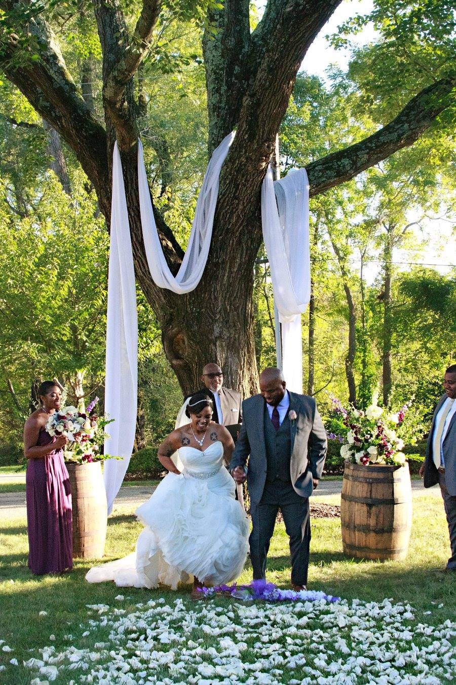 A wedding ceremony outdoors under a large tree decorated with white fabric. The bride and groom are walking together on flower petals on the grass, with a bridesmaid holding a bouquet nearby and two groomsmen standing to the side. The scene is bright and lush with green trees in the background.