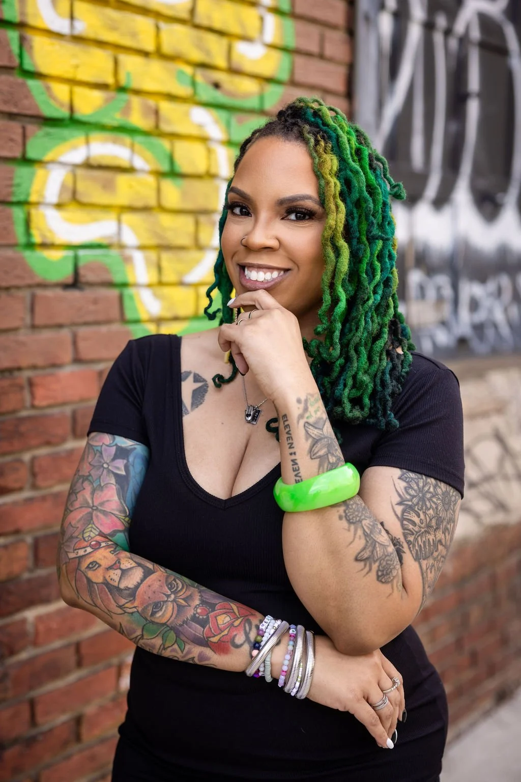 A woman with long, green and black dreadlocks, tattoos on her arms, wearing a black shirt, and smiling while standing in front of a brick wall with graffiti.