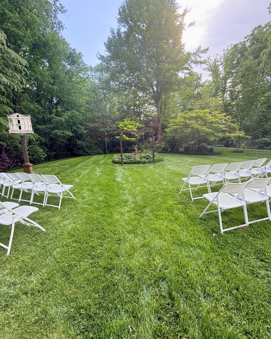 A backyard scene prepared for an outdoor event with lime green grass, rows of white chairs facing a garden arch, a birdhouse on a post, and tall trees surrounding the area.