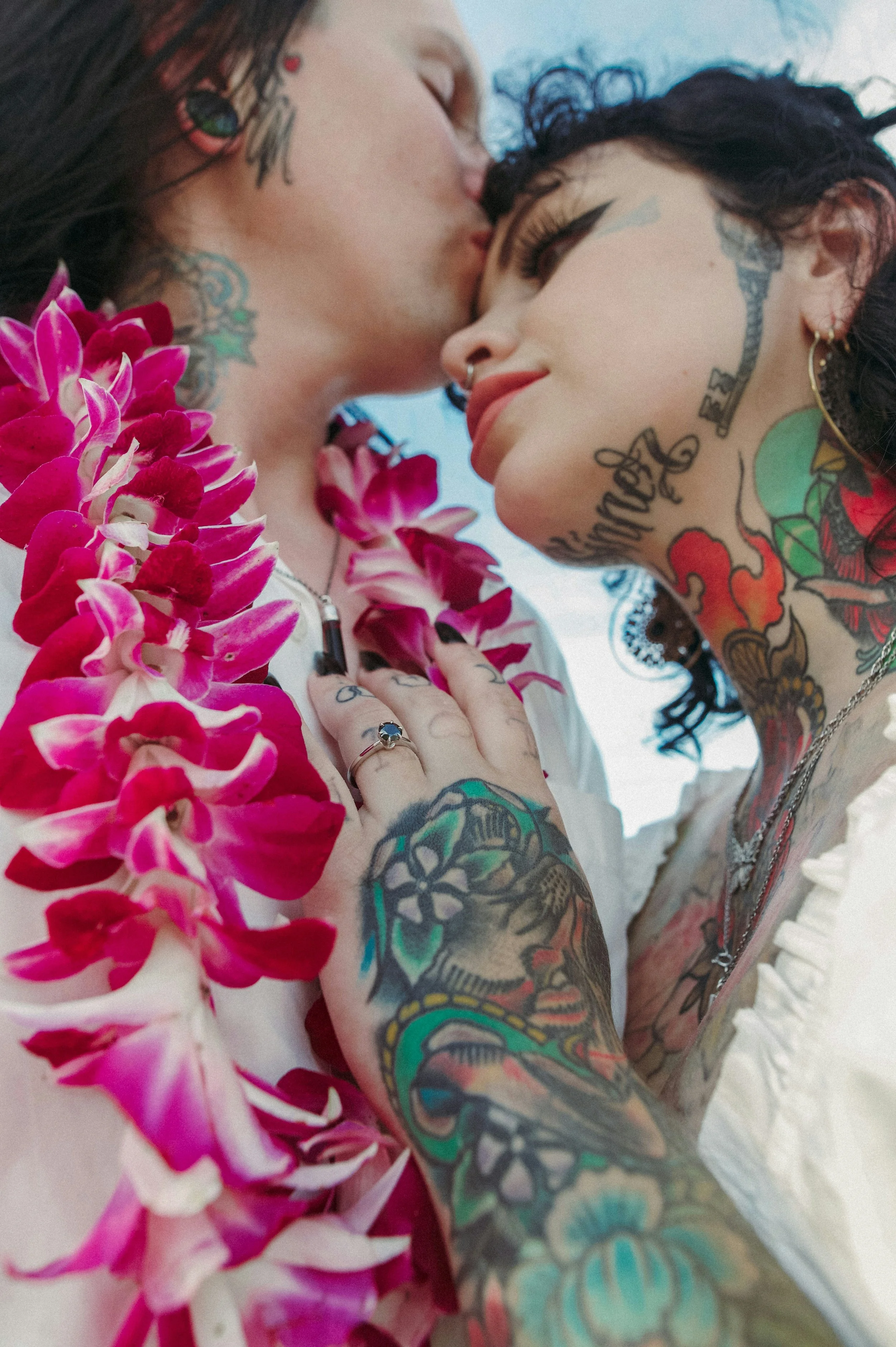 Two women with tattoos on their arms and necks share a close, intimate moment, touching noses and leaning in. They are near a cluster of vibrant pink flowers and are smiling softly.