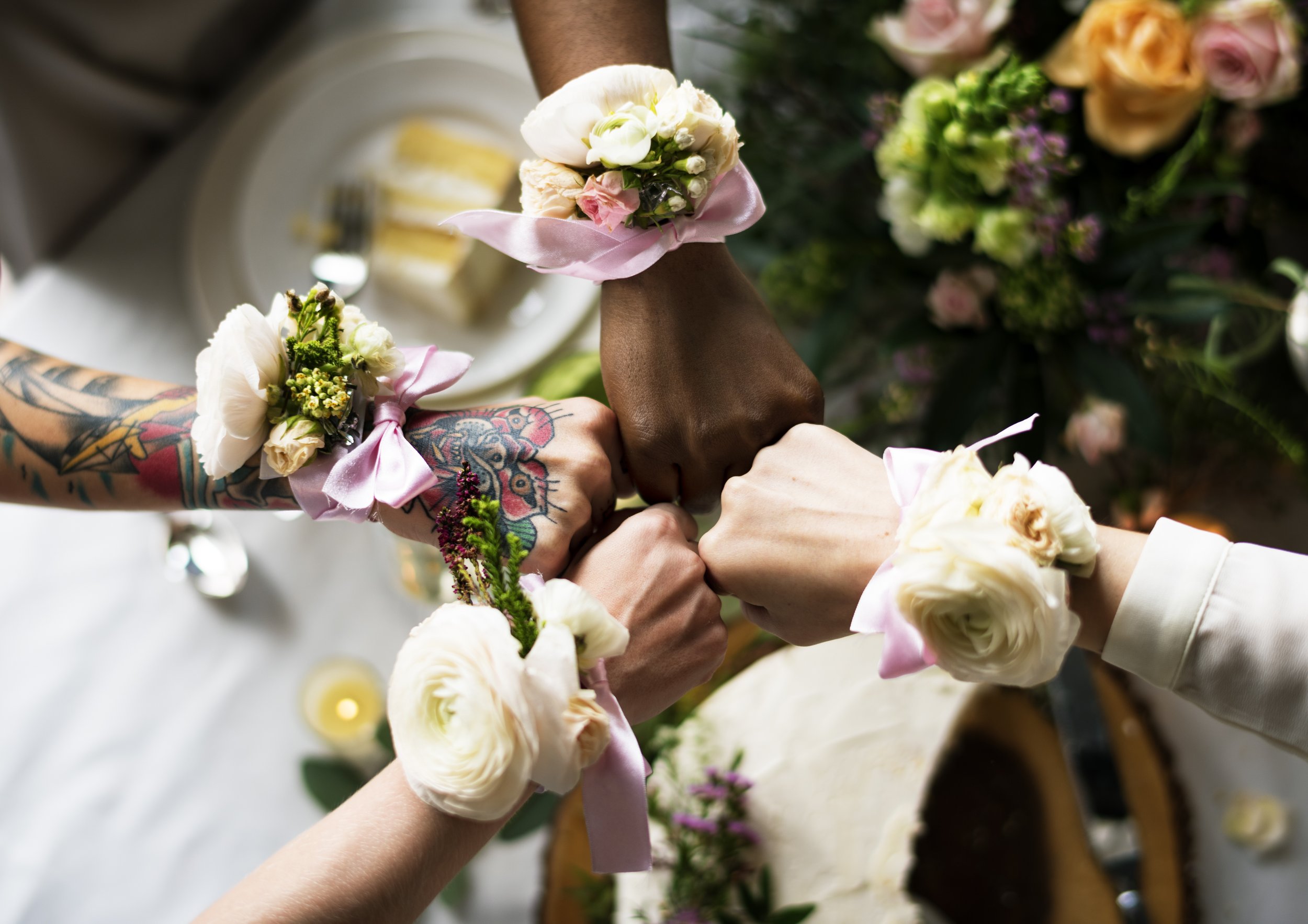 Close-up of multiple people holding hands during a wedding ceremony, each wearing floral wrist corsages made of white and pink roses secured with pink ribbons.