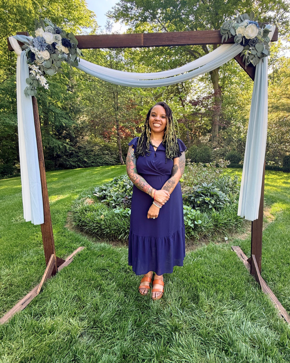 A woman with green dreadlocks and tattoos standing under a decorated wooden arch with white drapery and floral arrangements, outdoors in a green garden.