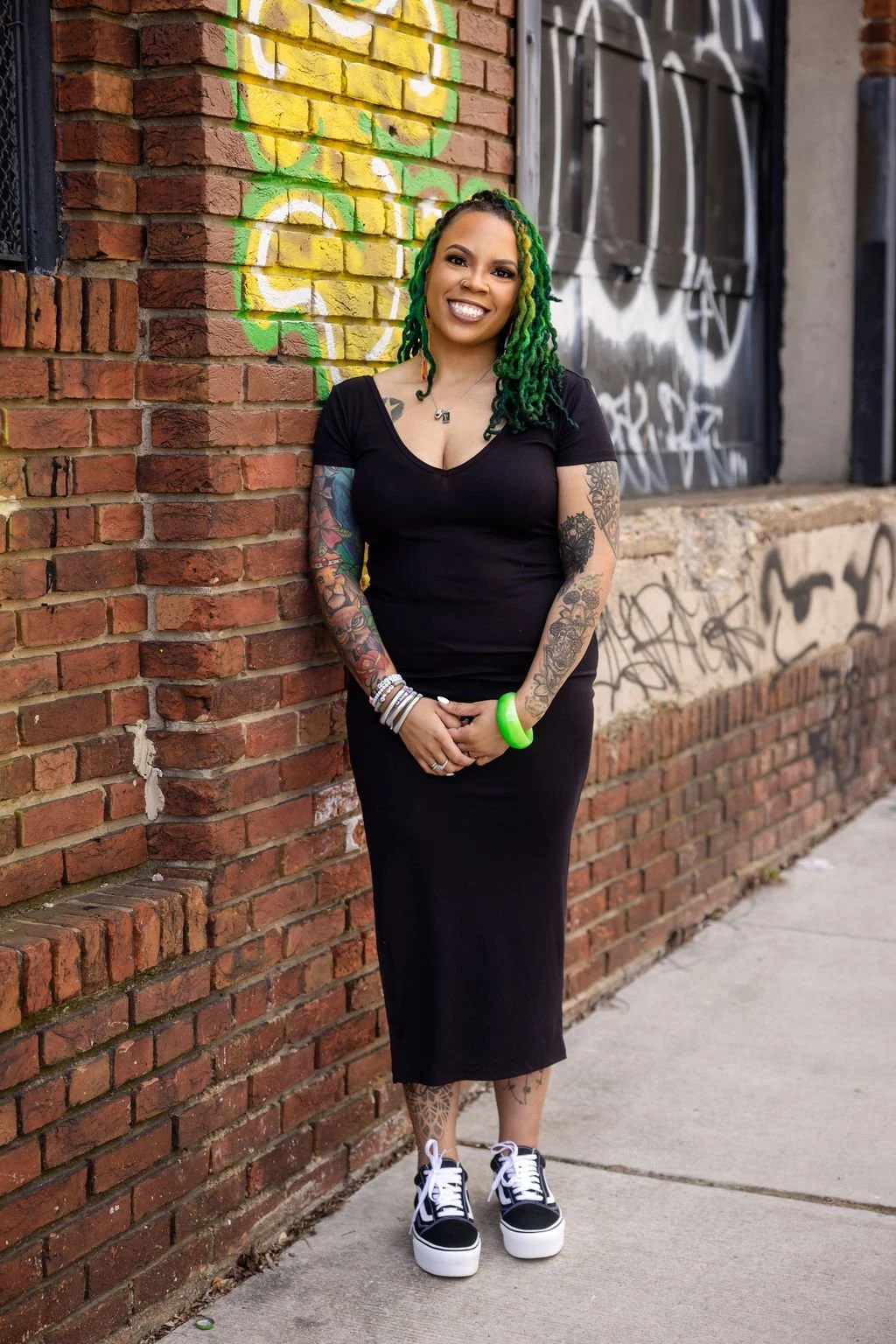 A woman with green dreadlocks, tattoos on her arms and legs, wearing a black dress, white and black sneakers, and green bracelet, standing against a brick wall with colorful graffiti.