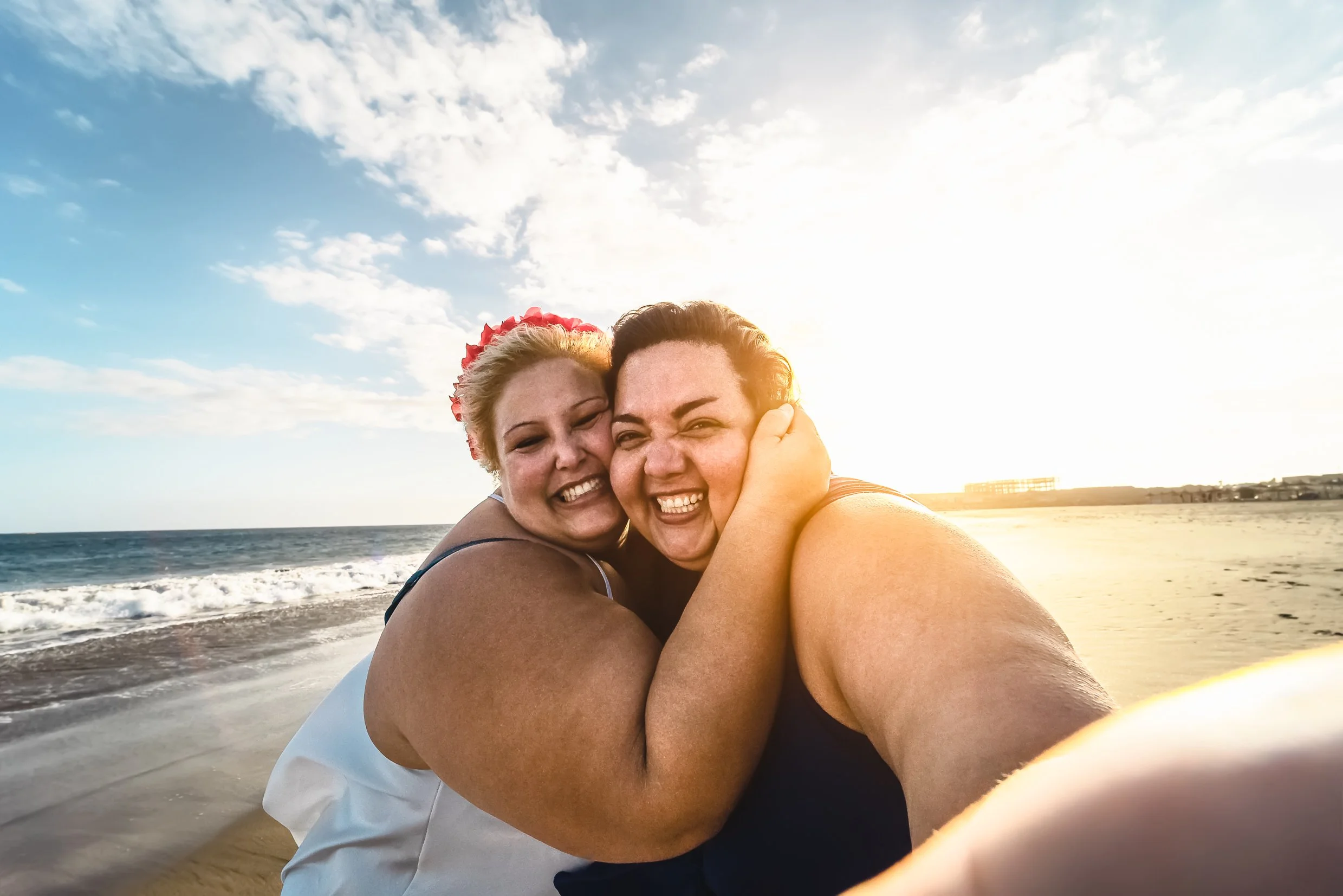 Two women hugging and smiling at the beach with the ocean, sand, and a partly cloudy sky in the background, during sunset.