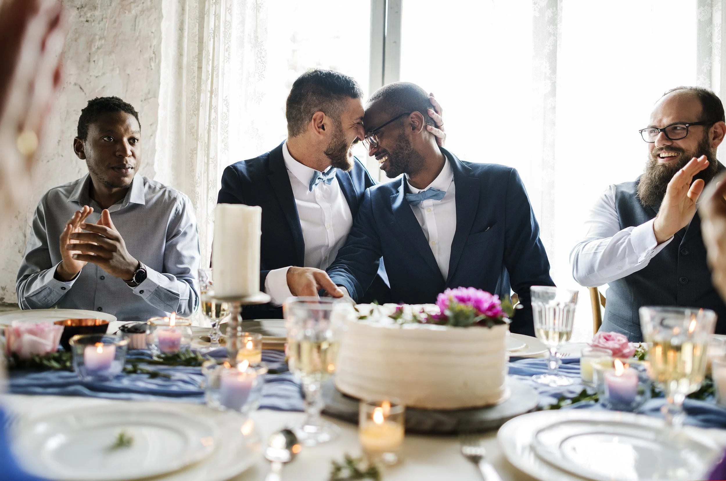 Four men dressed in suits and bow ties sitting at a decorated table during a celebration. Two men in the center are touching foreheads and smiling. The table has candles, plates, and floral decorations. Bright natural light is coming through lace curtains behind them.