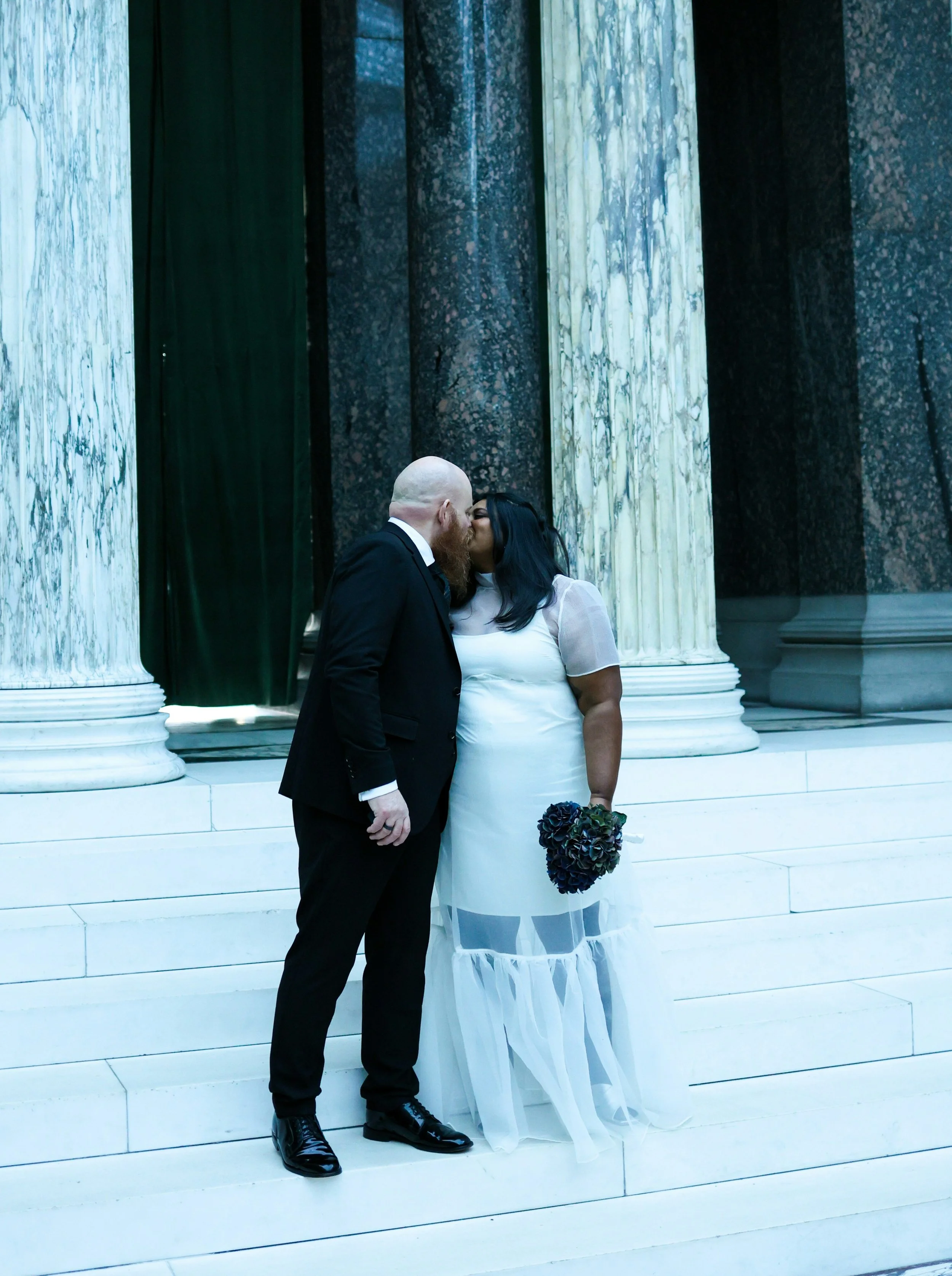 A couple dressed in formal wedding attire sharing a kiss in front of large marble pillars at a grand building.