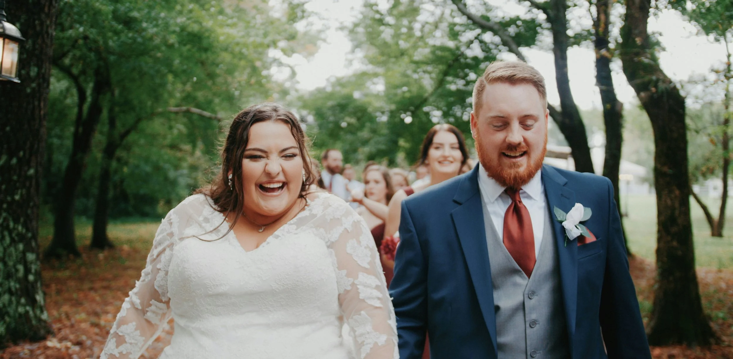 A bride and groom walking together outdoors at a wedding, surrounded by smiling guests and green trees.