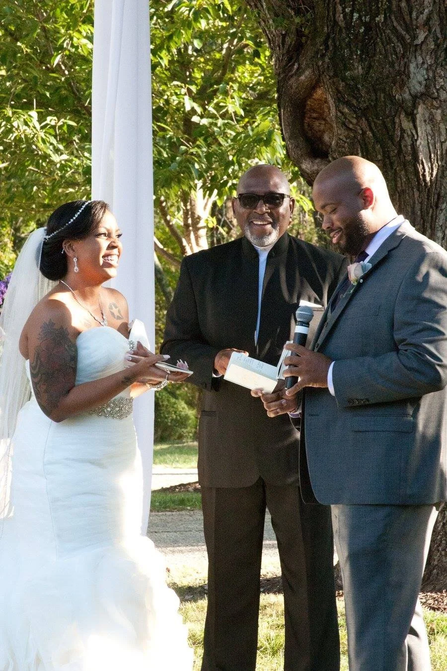 A bride and groom are standing outdoors during a wedding ceremony, smiling at each other. The officiant is holding a microphone and a book, and is dressed in black. The bride is wearing a white strapless wedding gown with tattoos on her arms, and the groom is in a gray suit. They are standing under a large tree with green foliage in the background.