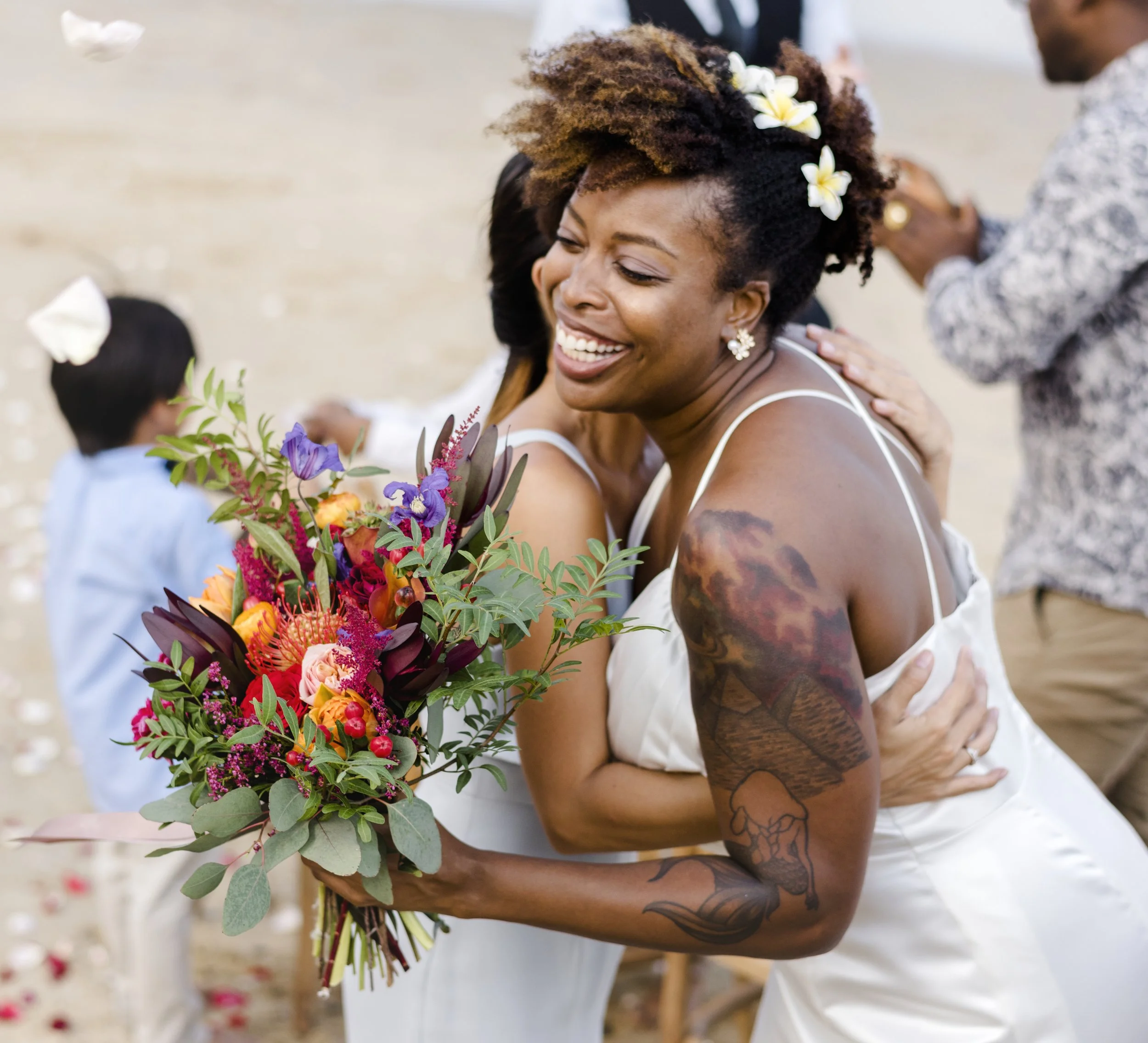 A woman with dark, curly, layered hair decorated with white flowers, wearing earrings, hugging someone at an outdoor beach wedding ceremony, holding a bouquet of colorful flowers. Part of her tattoos on her arm and shoulder are visible.