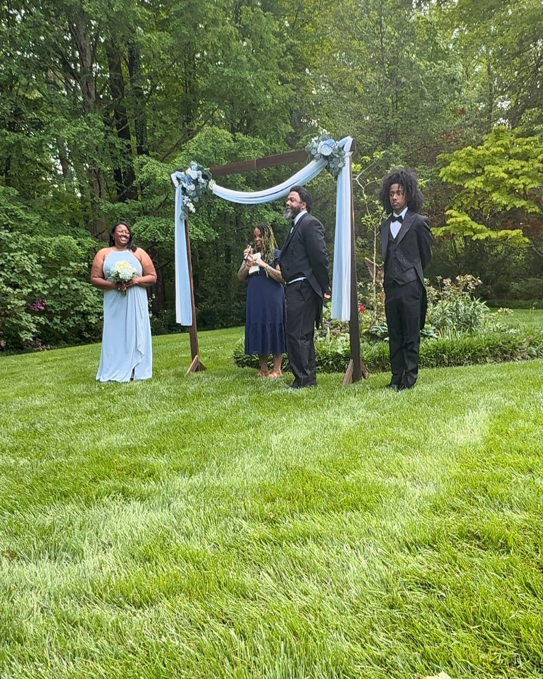A wedding ceremony outdoors under a decorated wooden arch with white and blue drapery and flowers, with three people in formal attire standing on a grassy lawn surrounded by trees.