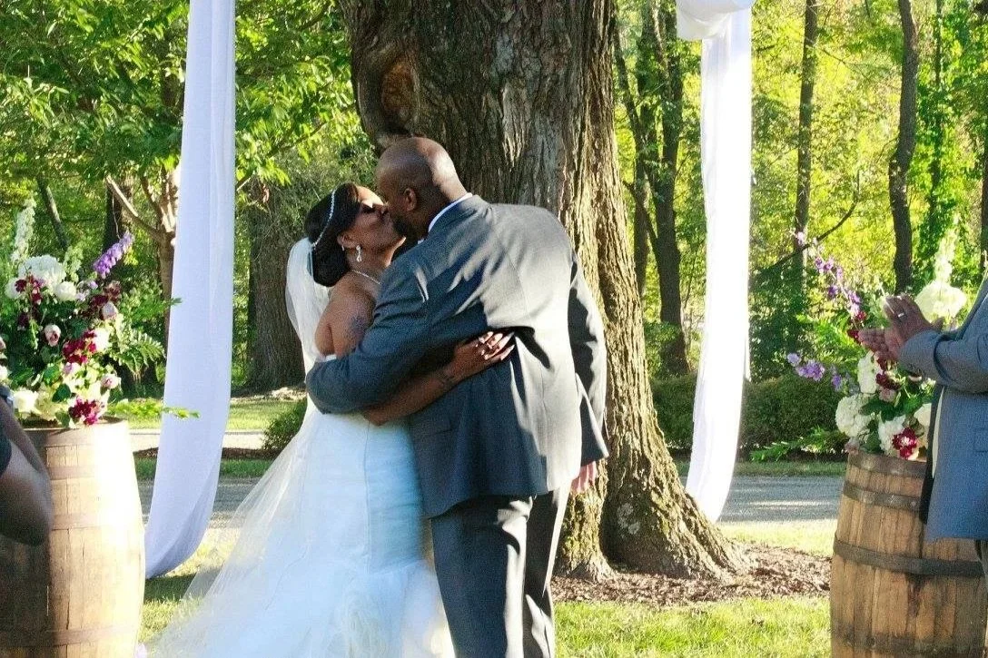 A bride and groom sharing a kiss during their outdoor wedding ceremony behind a tree, with floral arrangements and guests present.