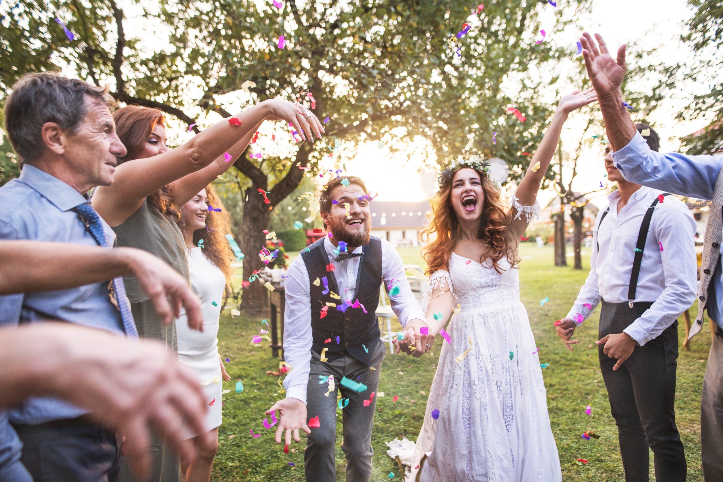 Group of people celebrating outdoors with confetti, smiling, and holding hands during sunset or late afternoon.