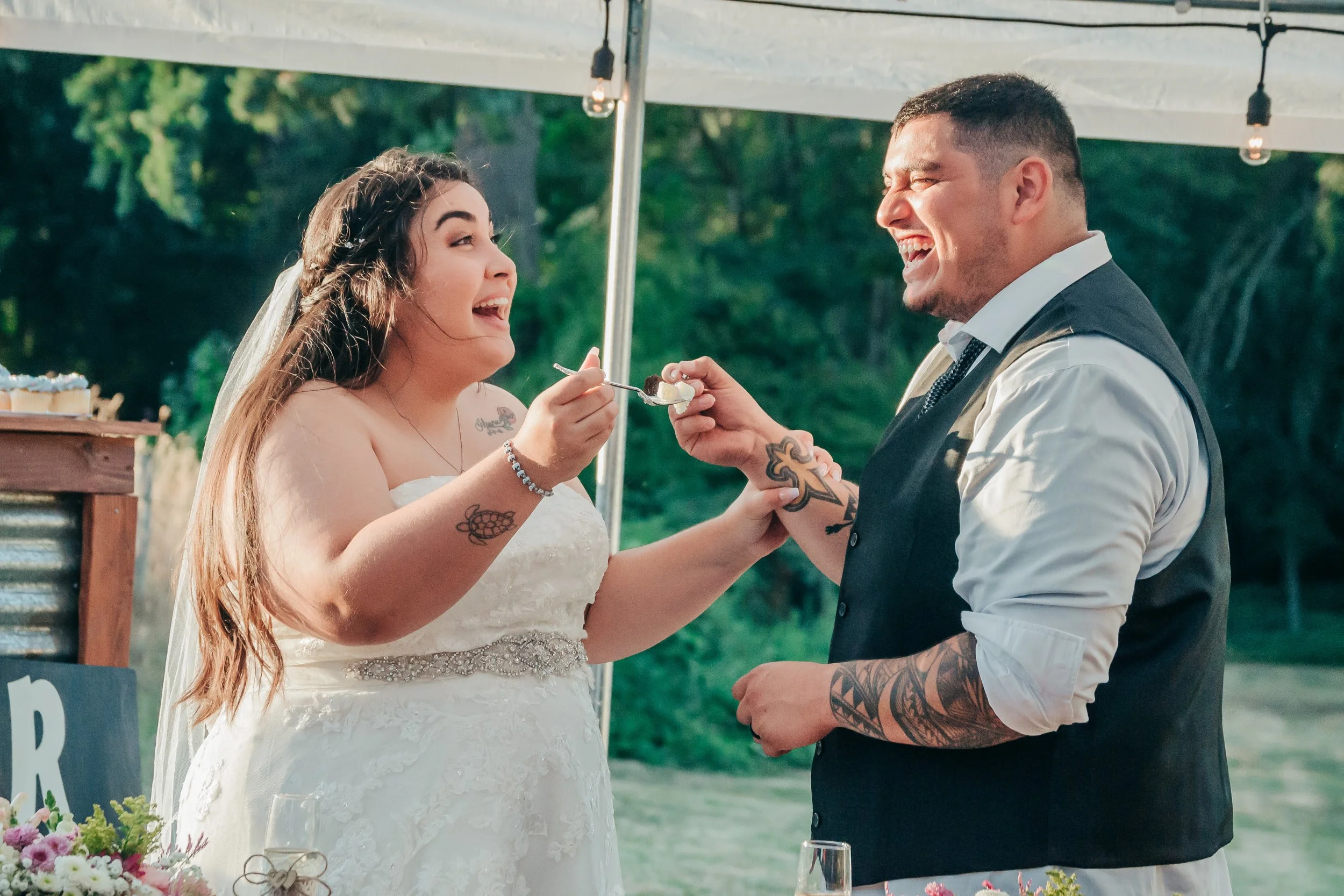 A bride and groom sharing a joyful moment at their wedding, with the bride wearing a white dress and the groom in a vest and white shirt, outdoors beneath a tent with string lights.