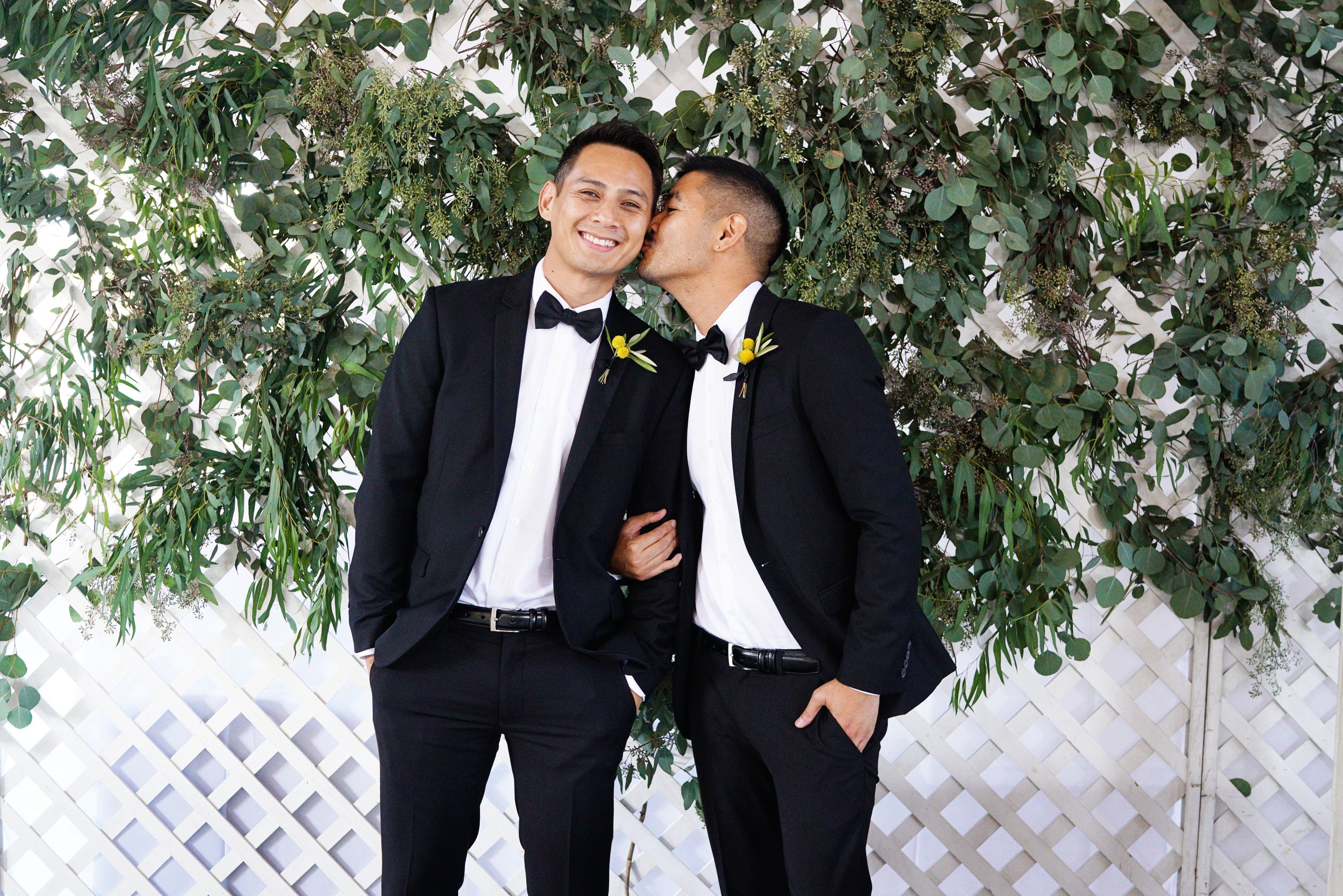 Two men in tuxedos, one kissing the other on the cheek, standing in front of a leafy green backdrop.