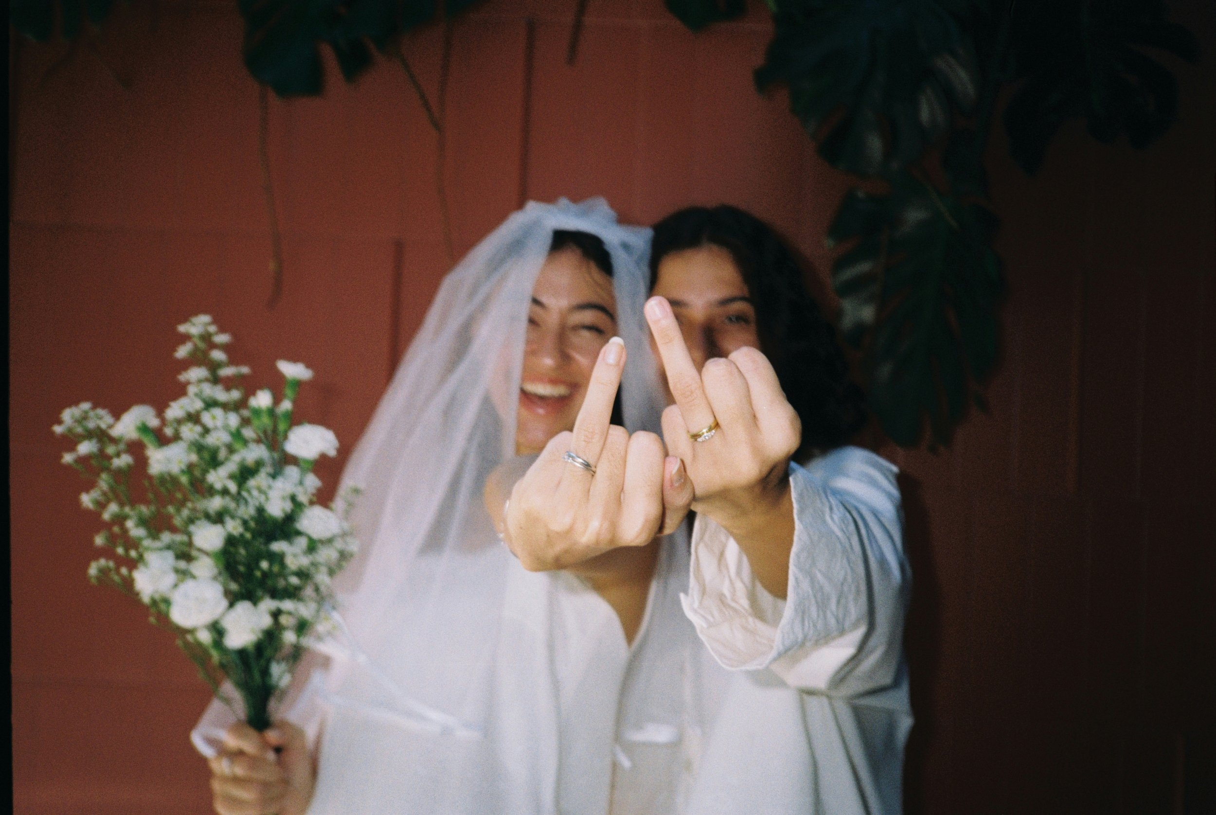 Two women, one wearing a wedding veil, smiling and making rude gestures with their middle fingers, holding a bouquet of flowers, possibly celebrating a wedding.