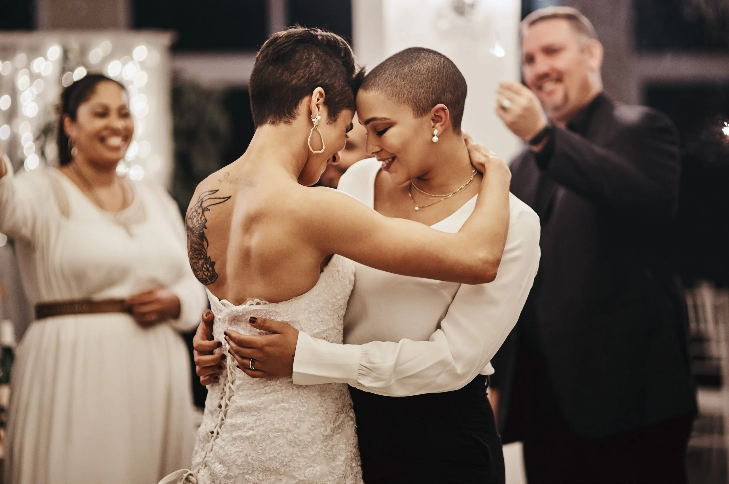 A couple wearing wedding attire dancing closely with their foreheads touching, surrounded by friends at a celebration.