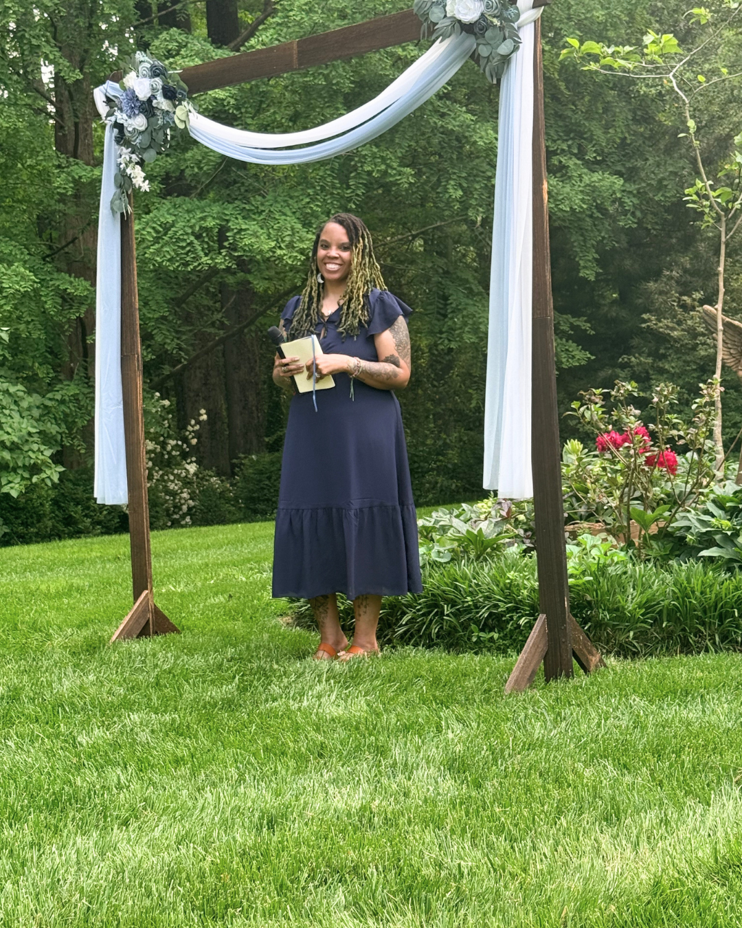 A woman with long dreadlocks, wearing a navy blue dress, stands under a wooden arch decorated with flowers and white draped fabric in a garden setting. She is smiling and holding a microphone and a small notebook.