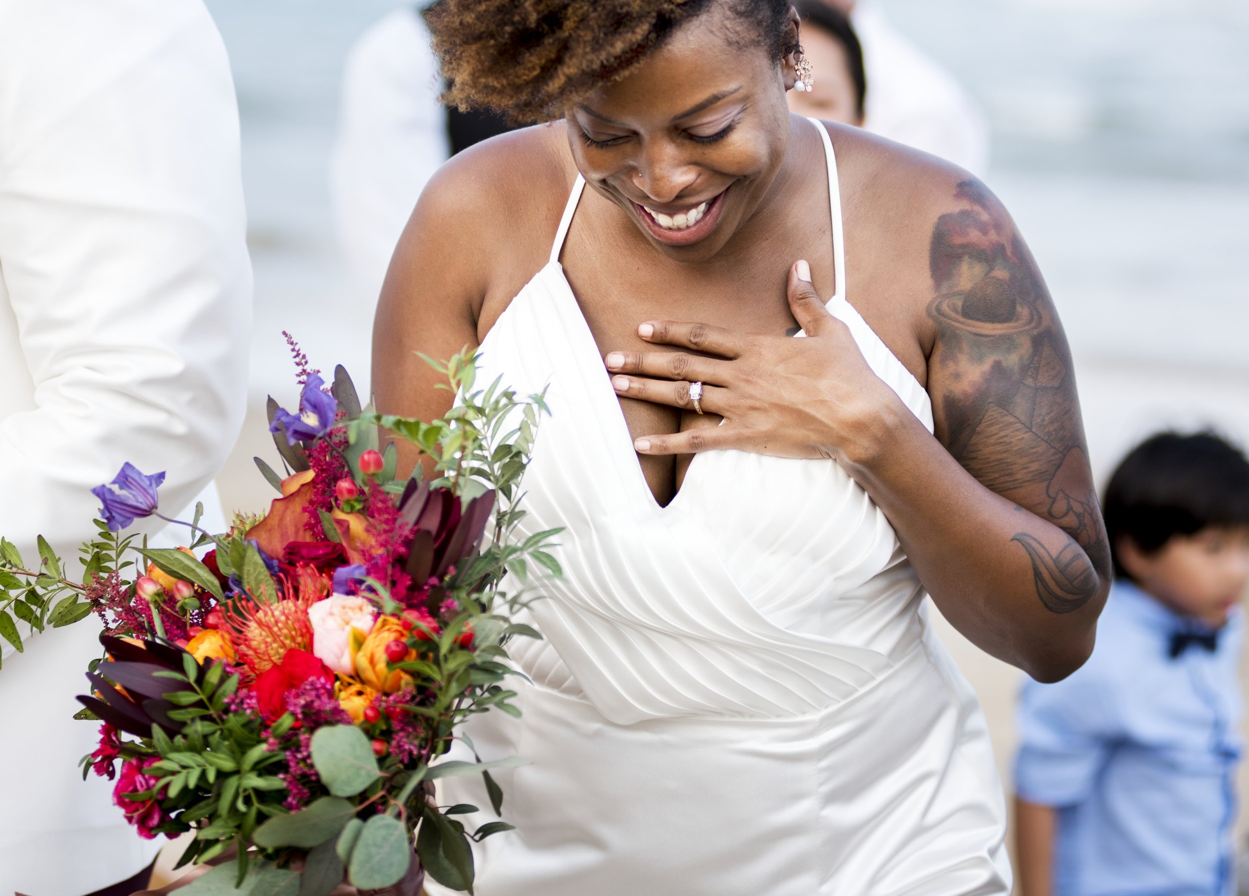 A woman in a white dress with a tattooed arm is smiling and placing her hand on her chest during a wedding ceremony. She is holding a large bouquet of colorful flowers.