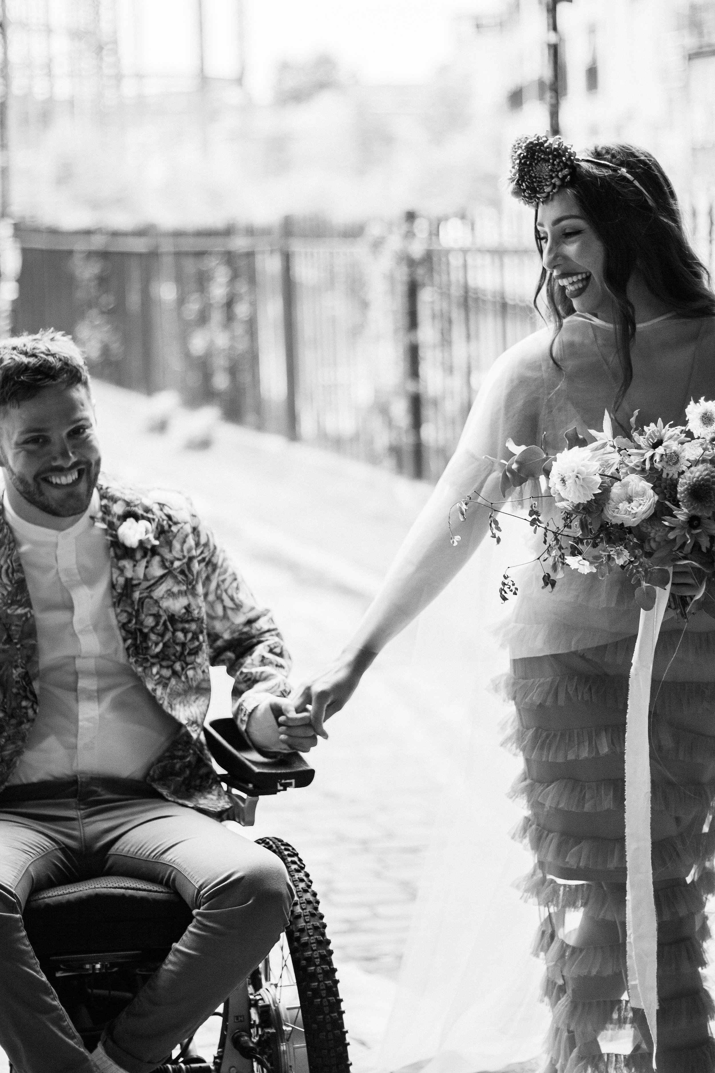 A woman in a ruffled dress and floral headpiece sharing a smile and holding hands with a man in a wheelchair, both outdoors in bright sunlight.