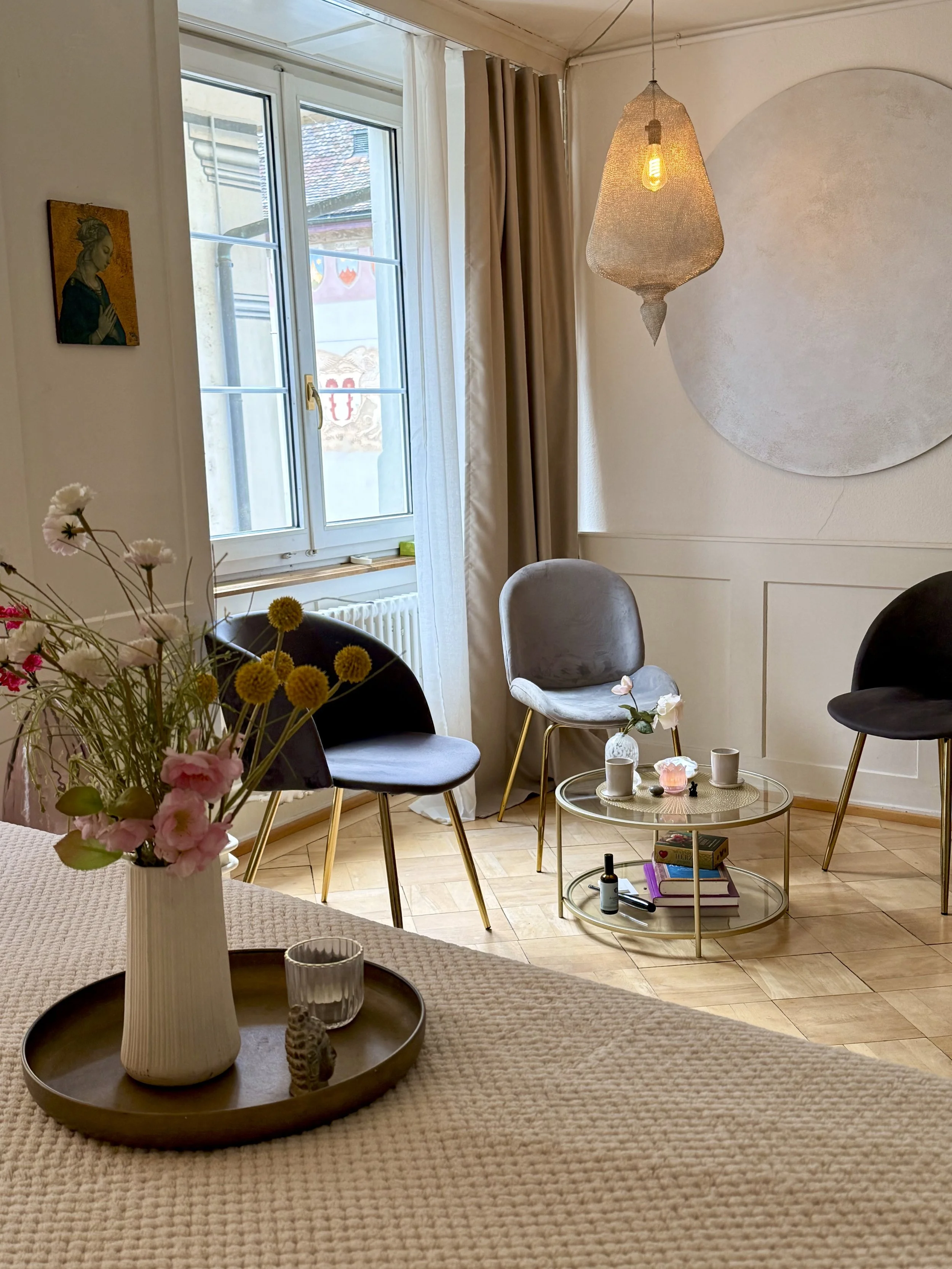 AYAMA Praxisraum in Rapperswil - Ein heller Raum mit großen Fenstern, einer Liege für Reiki Behandlungen, einem gemütlichen Sitzbereich mit drei Stühlen, einer dekorativen Wandlampe und einer runden Wanddekoration, alles in warmen, neutralen Farben.