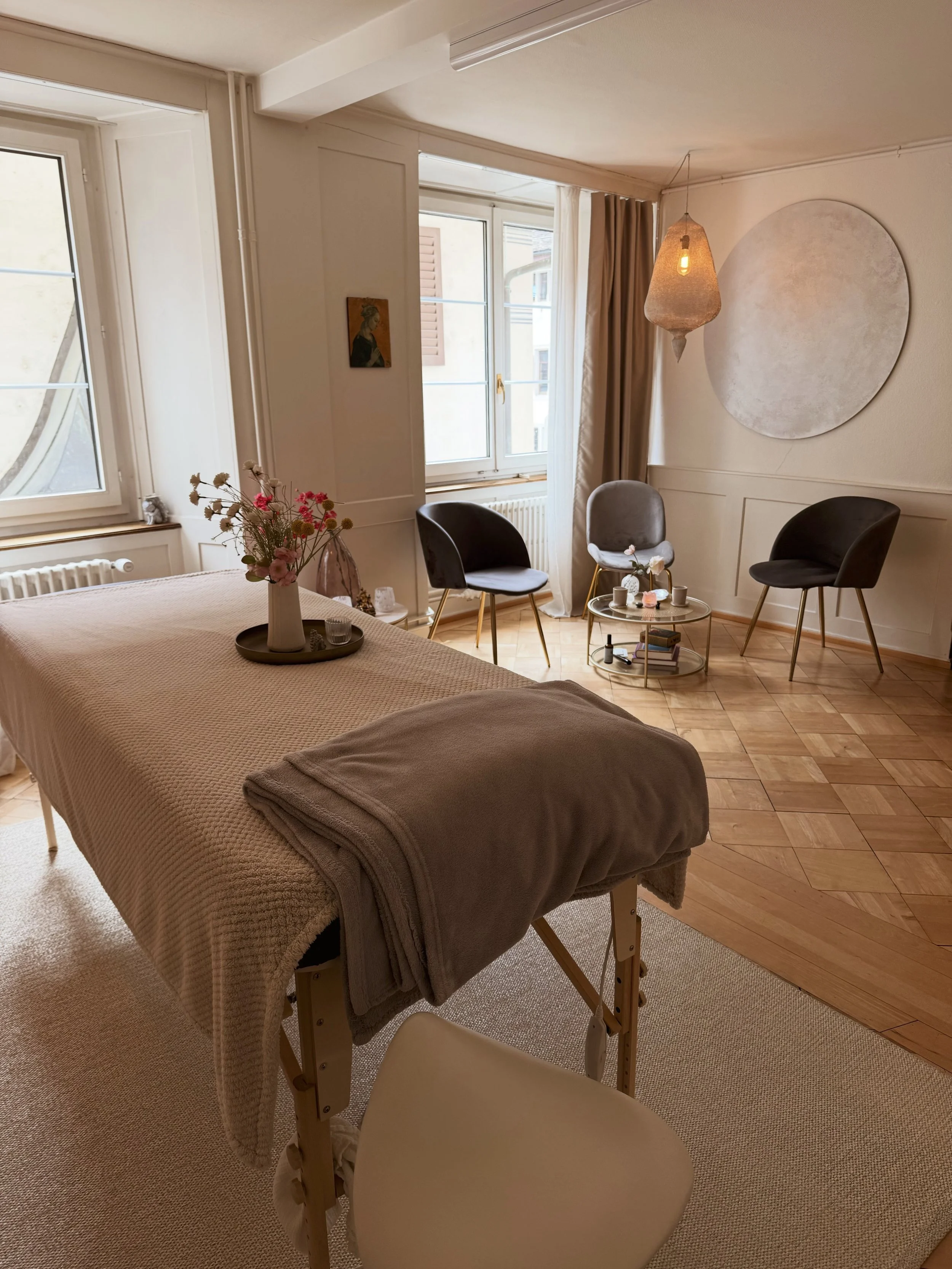 AYAMA Praxisraum in Rapperswil - Ein heller Raum mit großen Fenstern, einer Liege für Reiki Behandlungen, einem gemütlichen Sitzbereich mit drei Stühlen, einer dekorativen Wandlampe und einer runden Wanddekoration, alles in warmen, neutralen Farben.