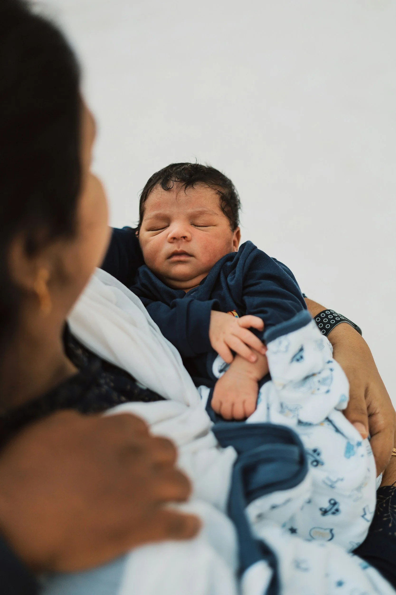A person holding a sleeping baby with curly hair in a dark blue outfit, against a plain white wall.