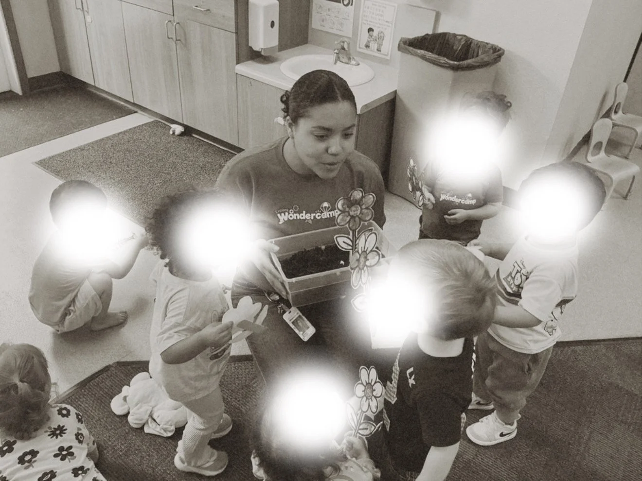 A woman surrounded by young children in a classroom, holding a flower-themed craft display and engaging with the children. The children are holding flower and paper cut-out crafts, and they are gathered on the floor and in front of the woman. The setting includes a sink, cabinets, a trash can, and children's chairs.